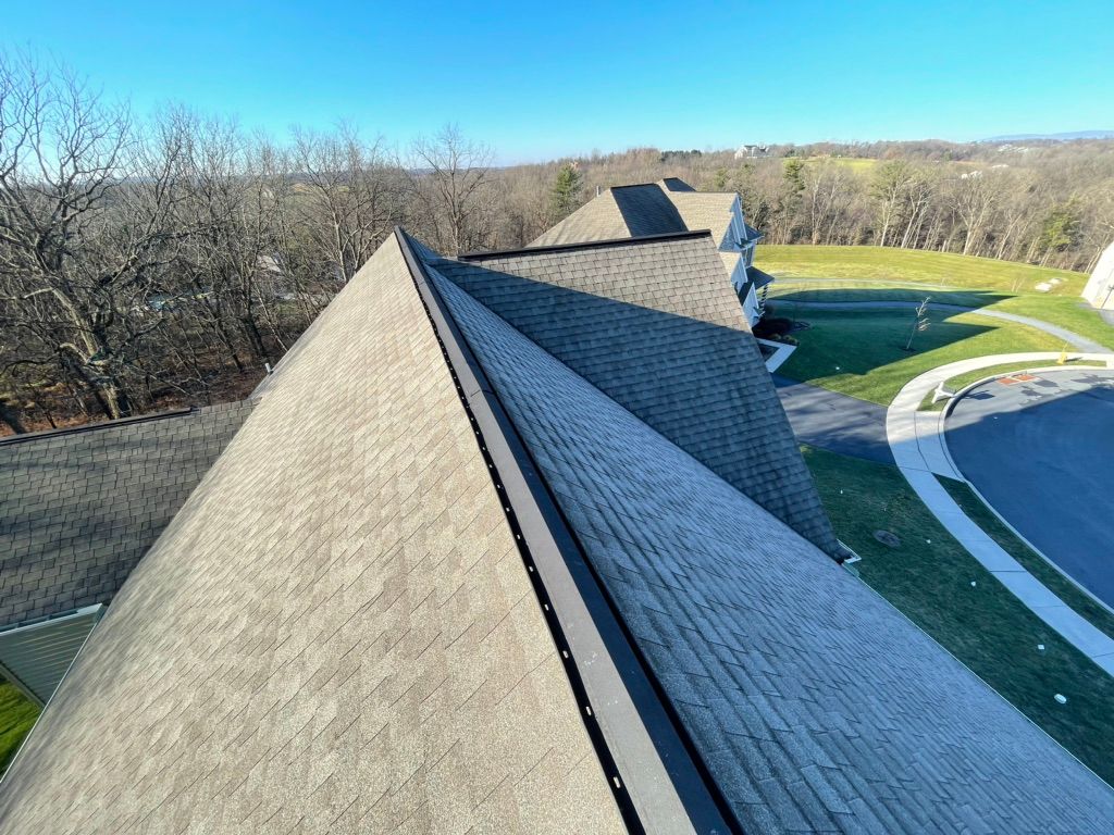 High-angle view of gray shingled residential roofs, a dark ridge vent, and a paved road leading to a grassy hill.