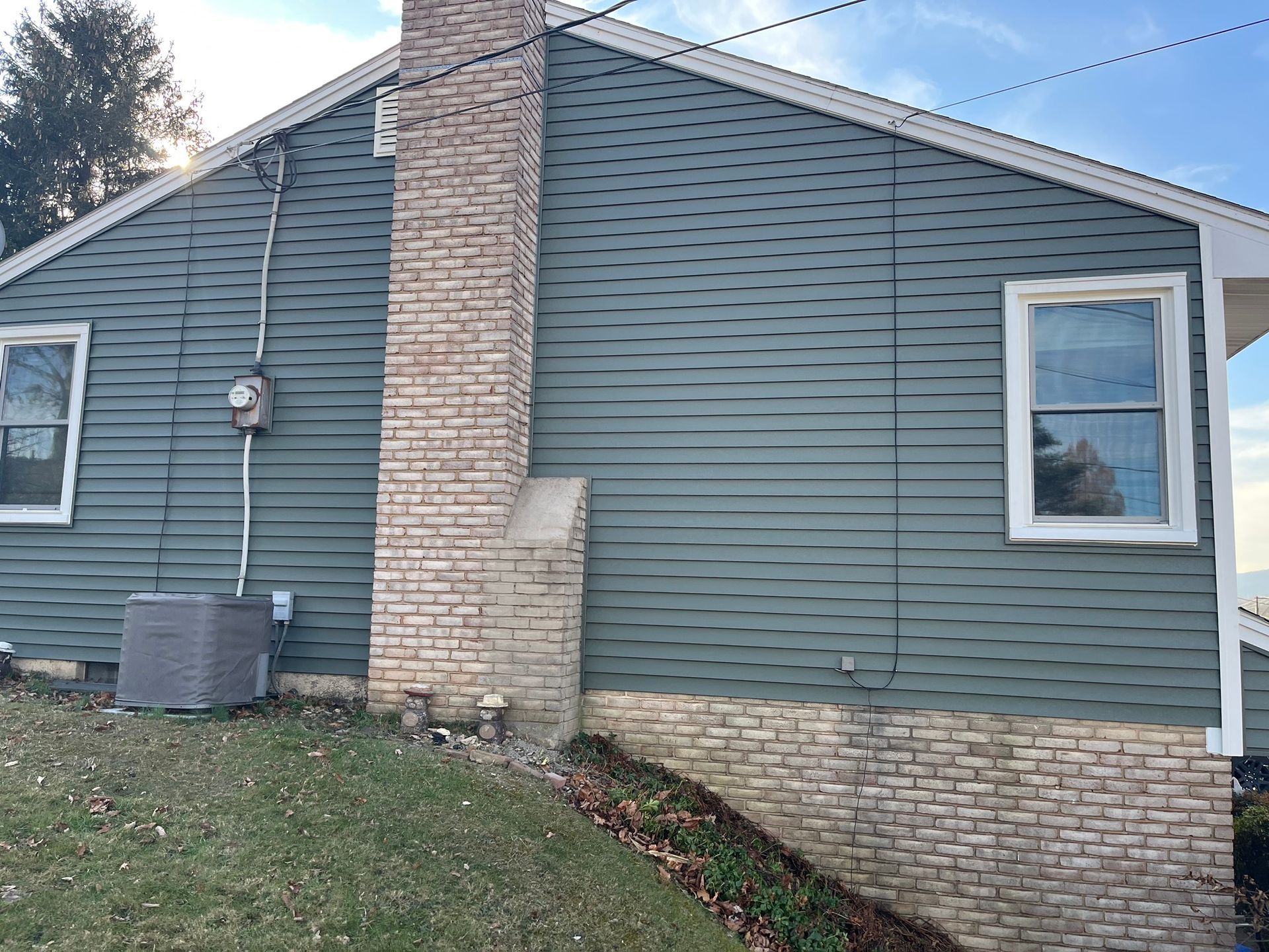 A side view of a house with dark gray horizontal siding, a light brick chimney, a stone foundation, and two windows.