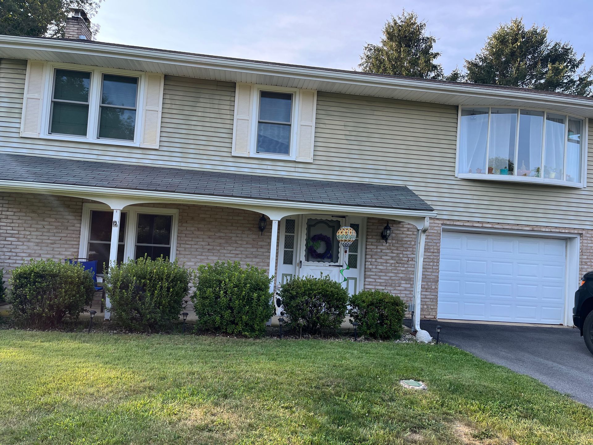 A beige, two-story house with a white garage, a covered front porch, and mature green shrubs in the front yard.