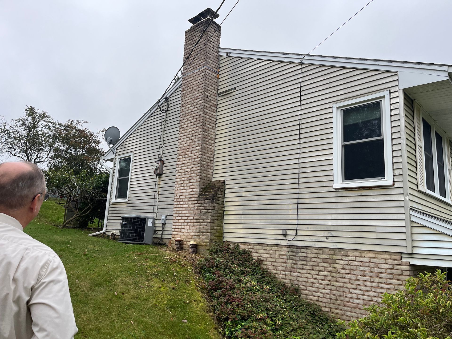 A person stands on a grassy lawn looking at the side of a house with vinyl siding, a brick chimney, and stone foundation.
