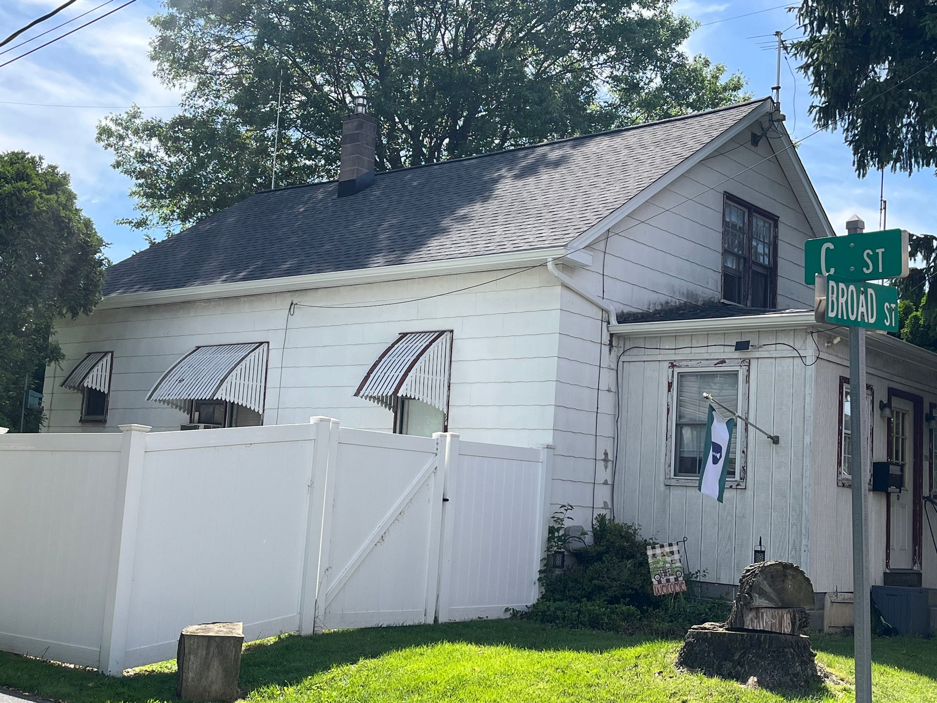A white, single-story house with a grey shingled roof, metal awnings, and a privacy fence on a sunny day.