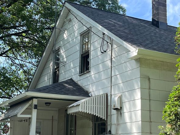 A two-story house with white siding, a shingled roof, a chimney, and a small front porch under a leafy tree.