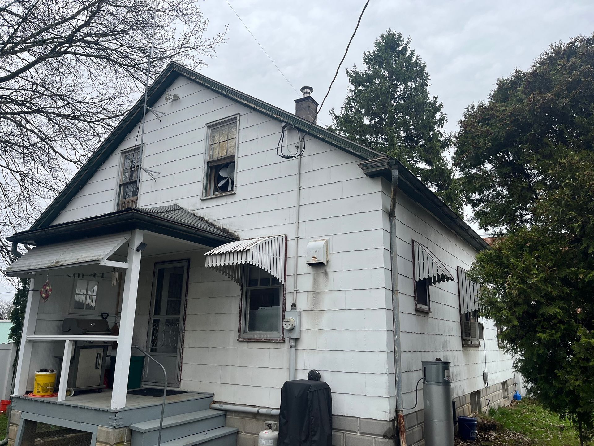 A weathered, two-story white house with a front porch, metal awnings, and visible exterior siding.