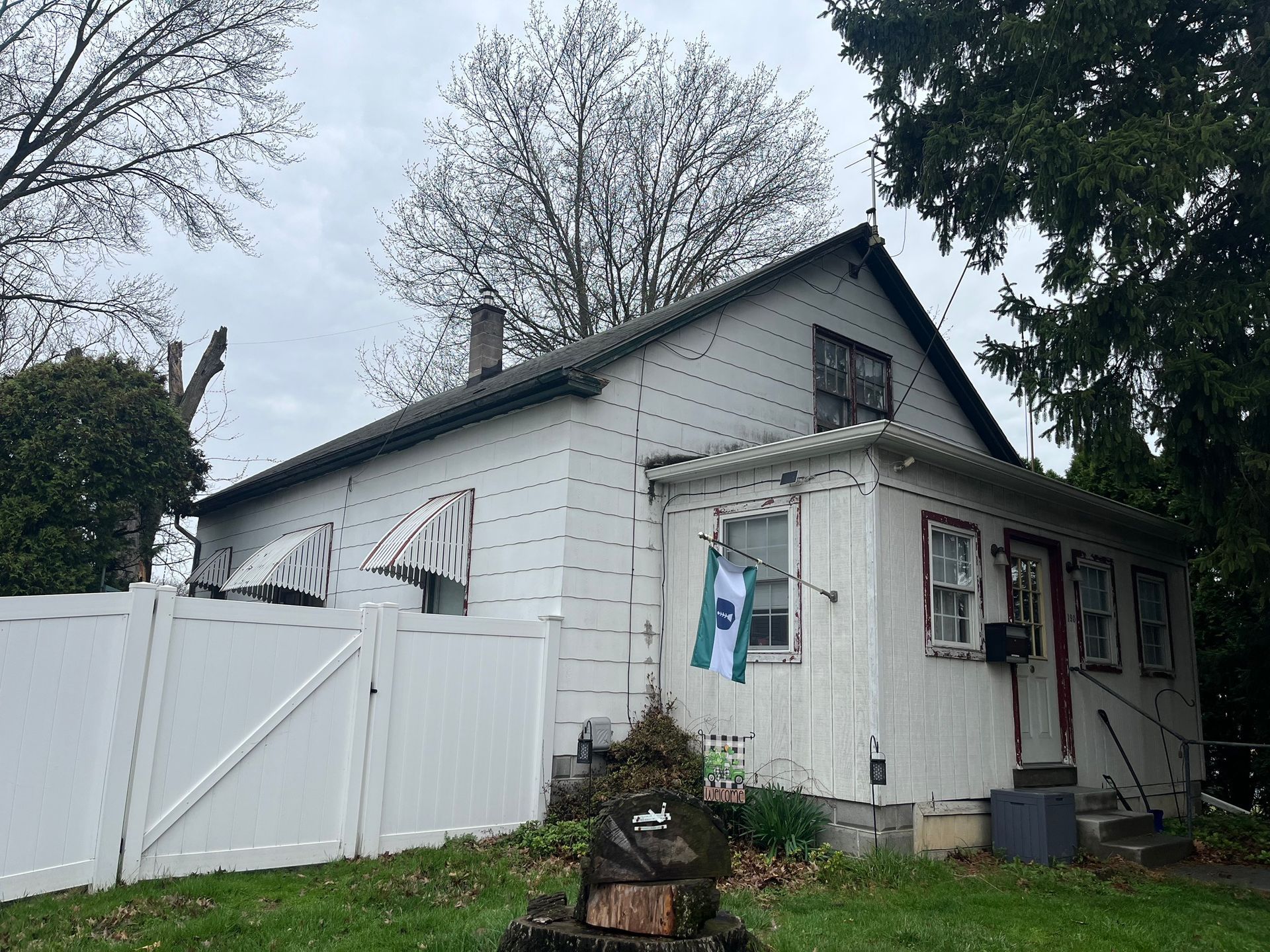 A white, one-story house with a peaked roof, small porch, and a white privacy fence in a yard with scattered trees.