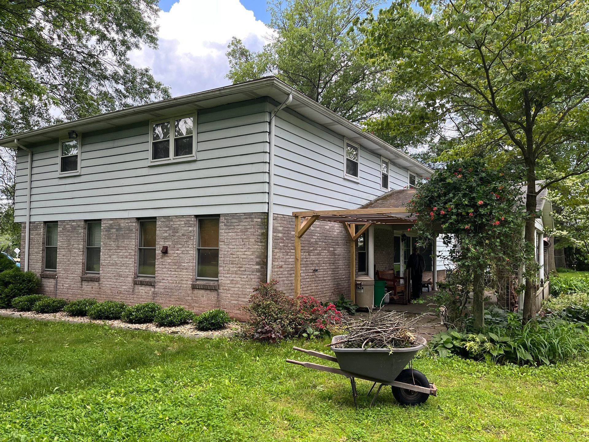 A two-story house with gray horizontal siding and a stone base sits in a lawn, with a wheelbarrow in the foreground.