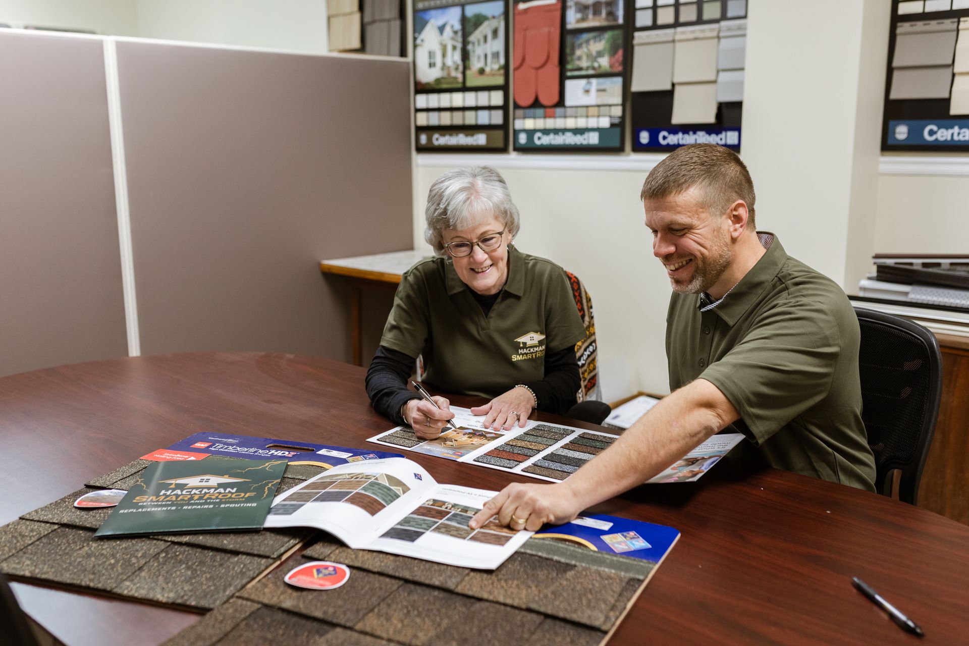 Two people in green shirts sit at a table reviewing roofing samples and brochures inside an office.