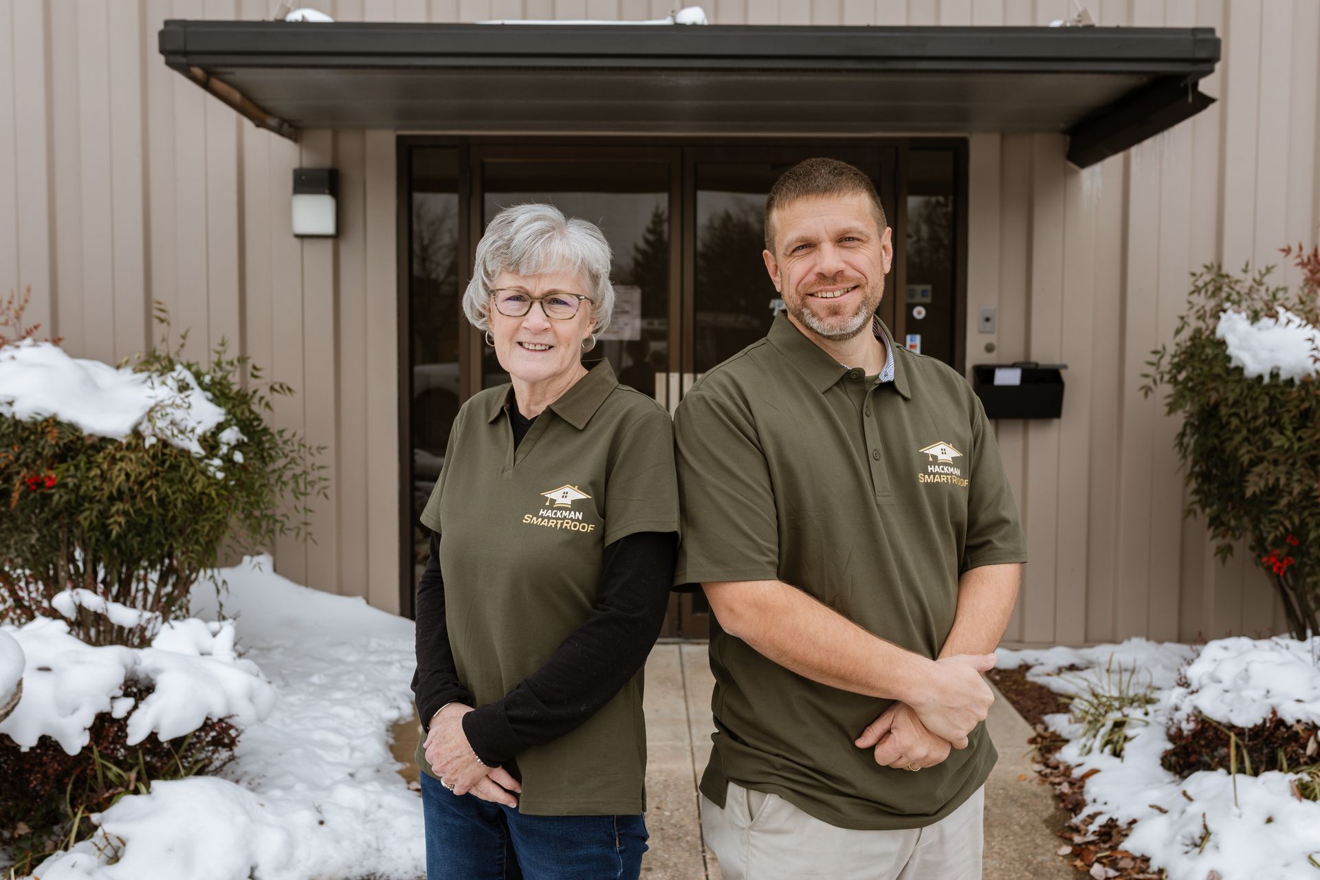 Two people wearing matching olive polo shirts stand smiling side-by-side in front of a building entrance with snow outside.