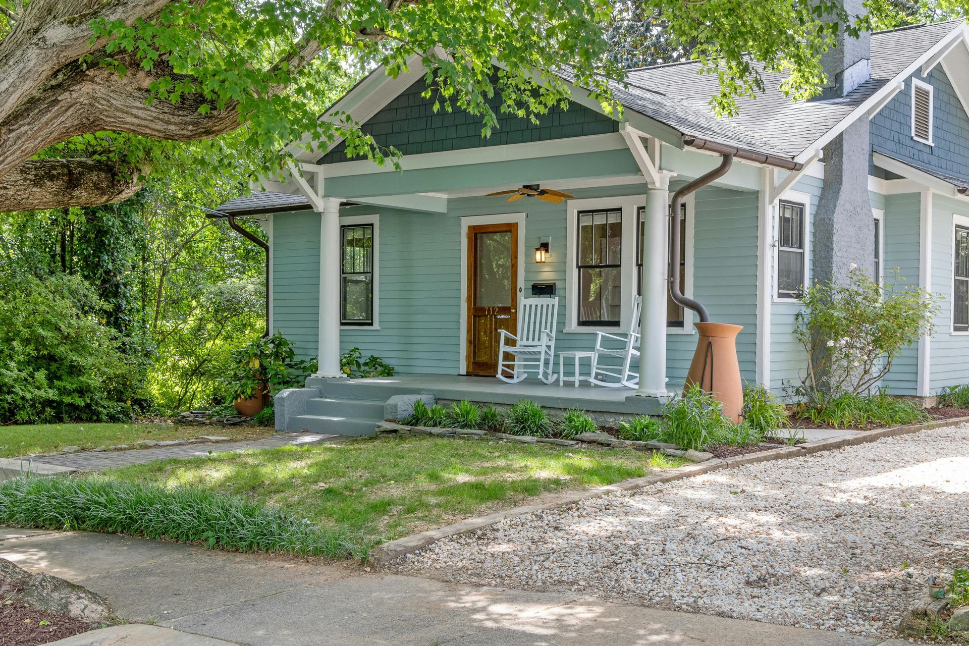 A light blue house with a covered front porch, white trim, and a gravel driveway, shaded by a large tree.