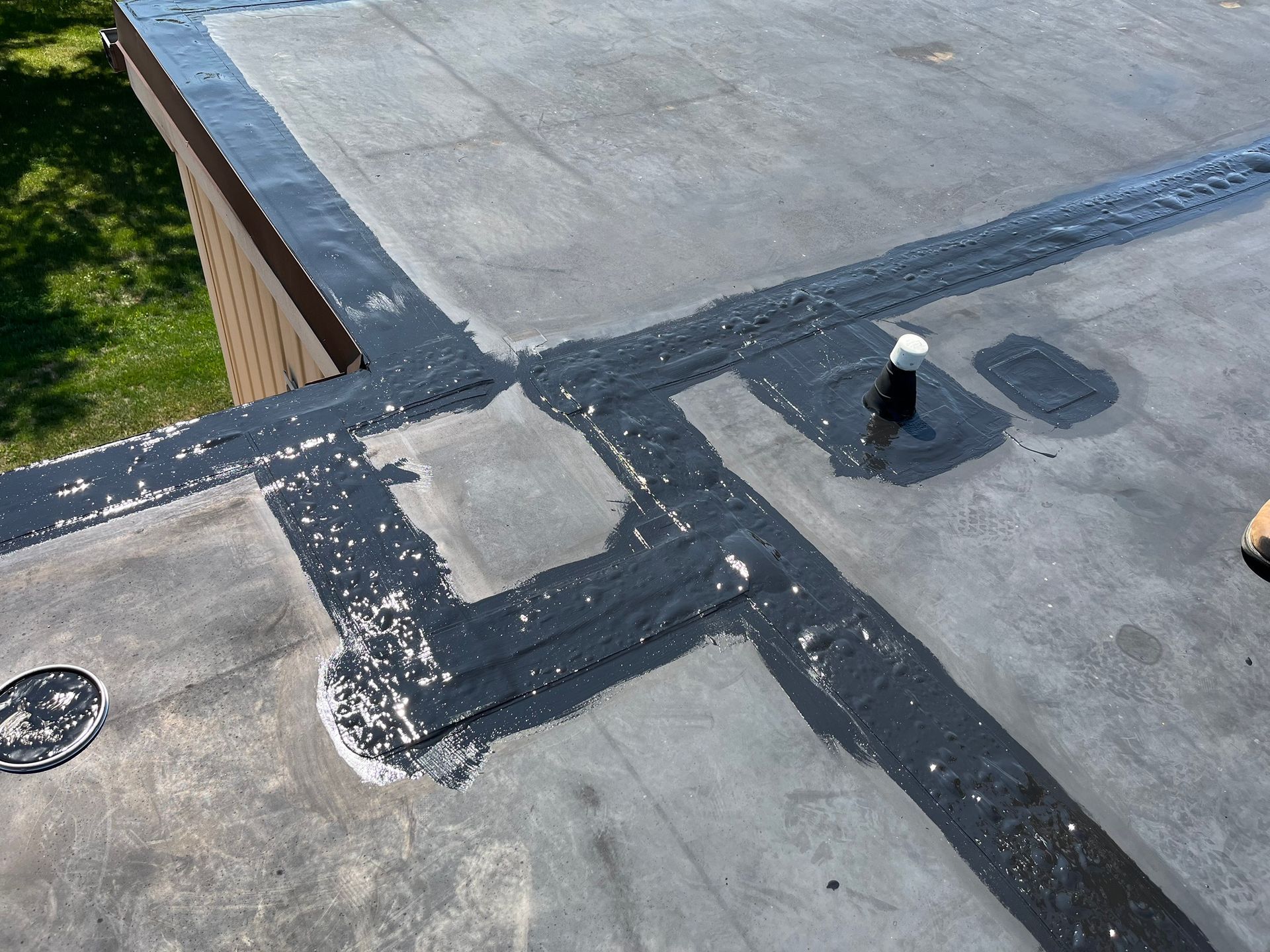 A gloved hand uses a handheld pressure roller to seal a seam on a black roofing membrane over wooden decking.