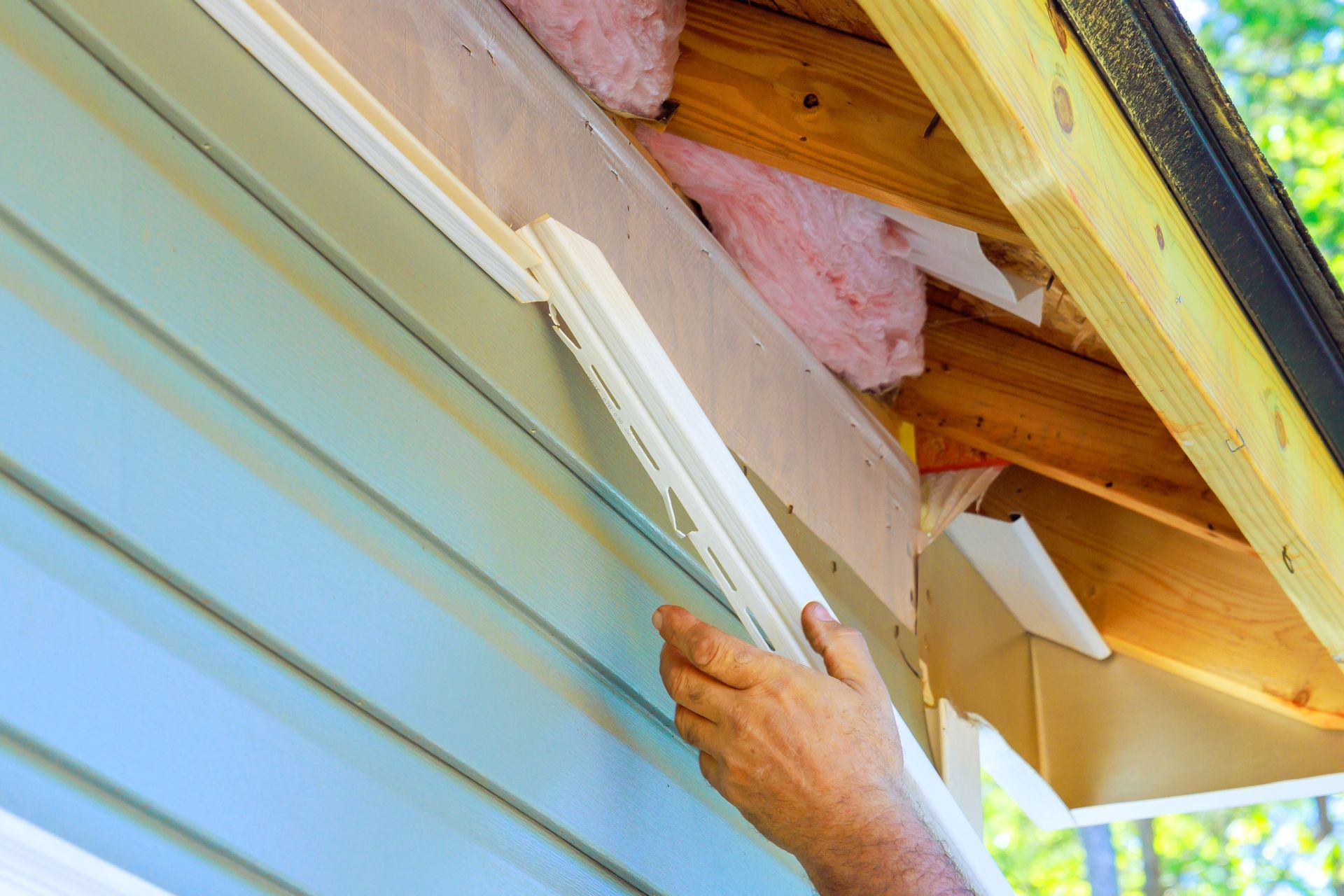 A person installing white vinyl siding trim along the edge of a house roof with pink insulation visible.