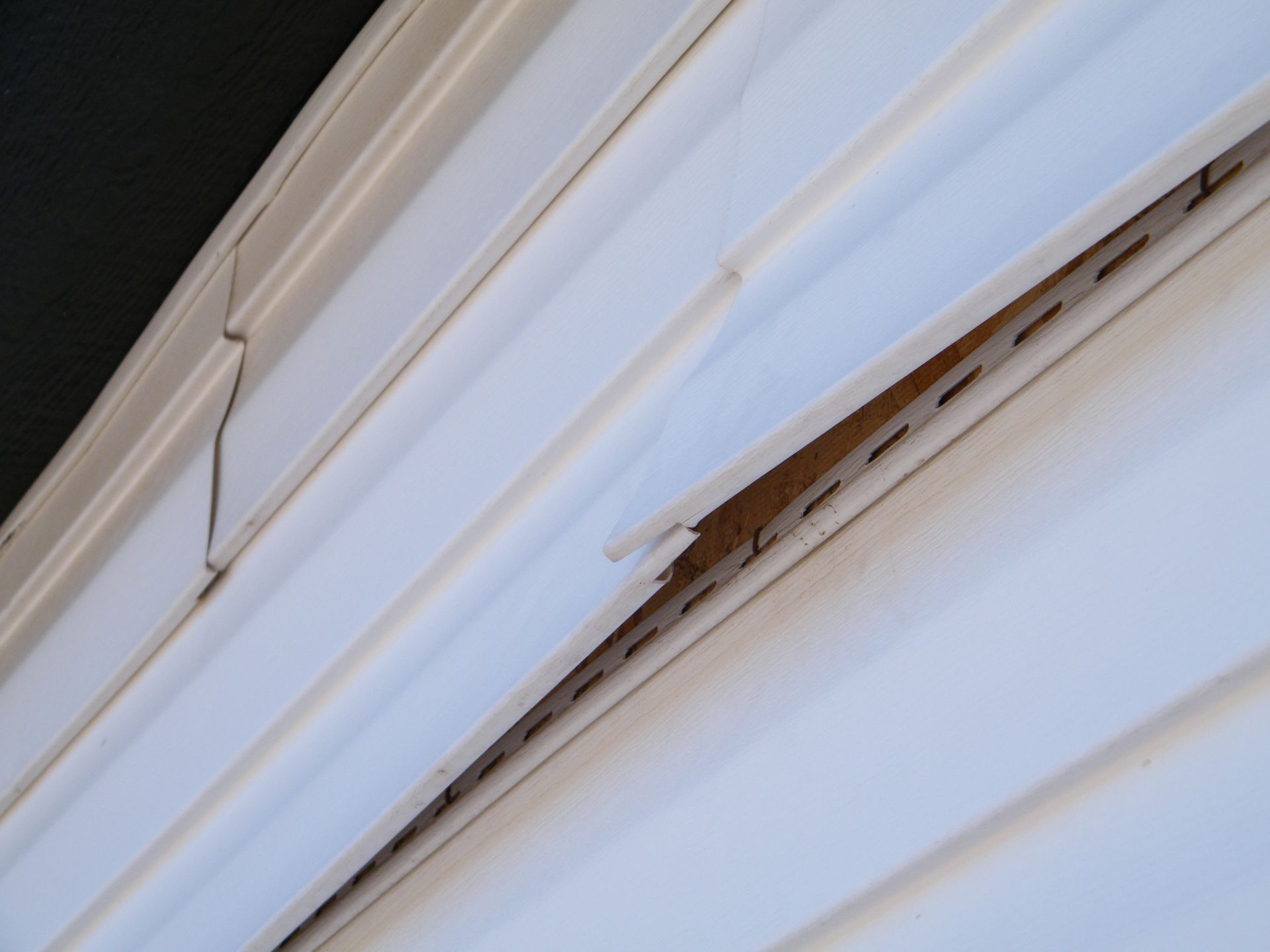 A close-up view of damaged white vinyl siding on a building exterior, showing a cracked and detached panel.