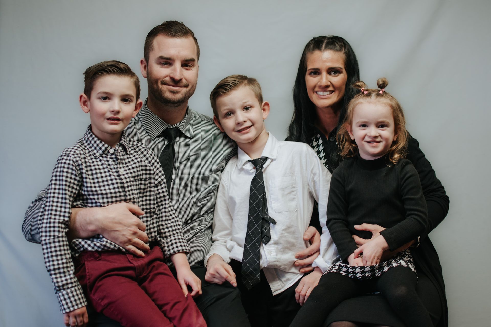 Family portrait: man, woman, and three children smiling, formally dressed, against a gray backdrop.