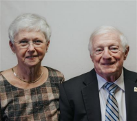 An elderly woman and man smiling side-by-side. Woman has short white hair, glasses and a patterned top. Man in a suit.