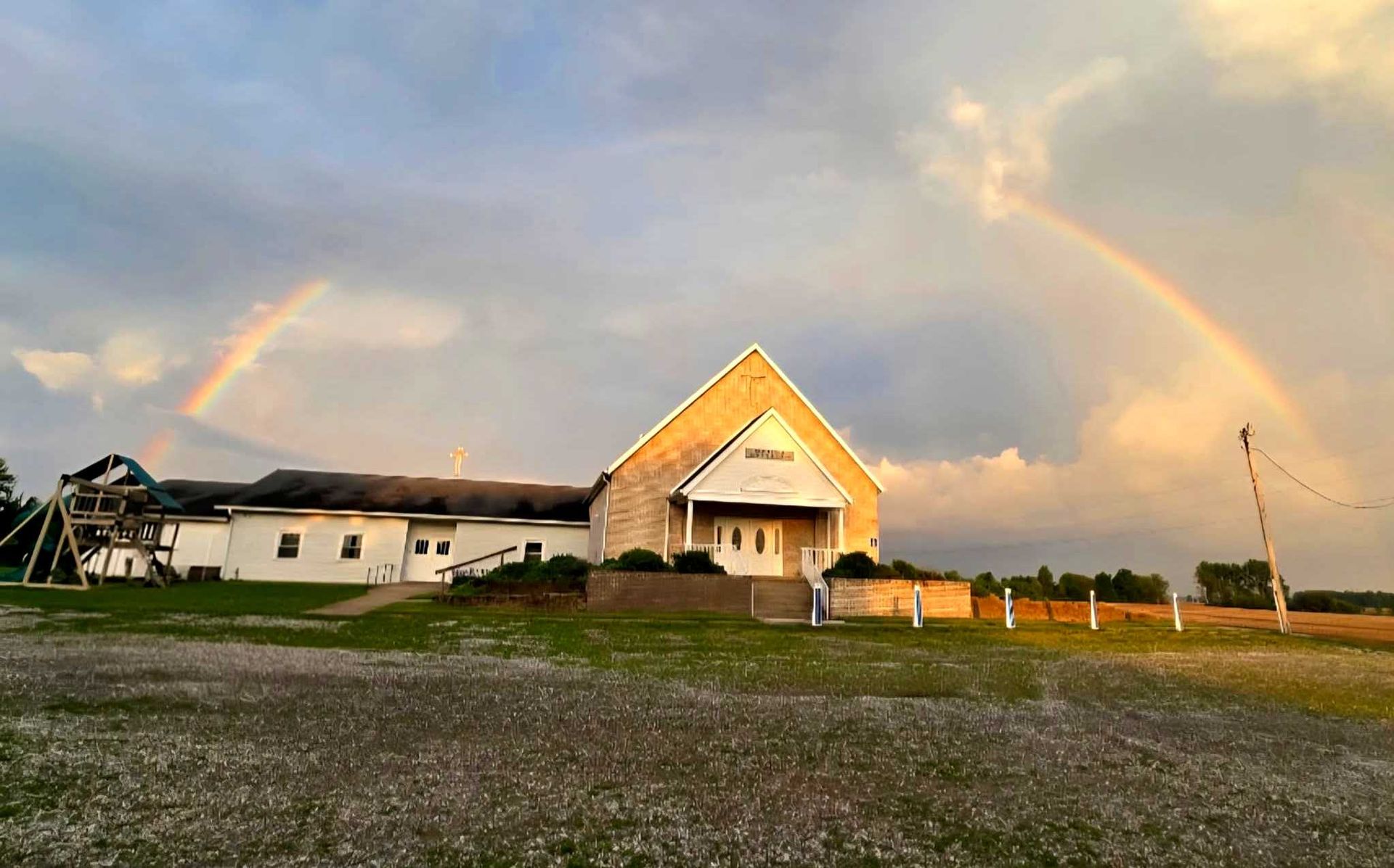 Church building with a rainbow arching over it in a field; sunset colors.