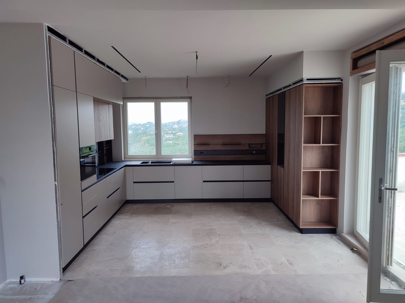 Cucina moderna con mobili grigio chiaro, dettagli in legno e una finestra con vista sul paesaggio.