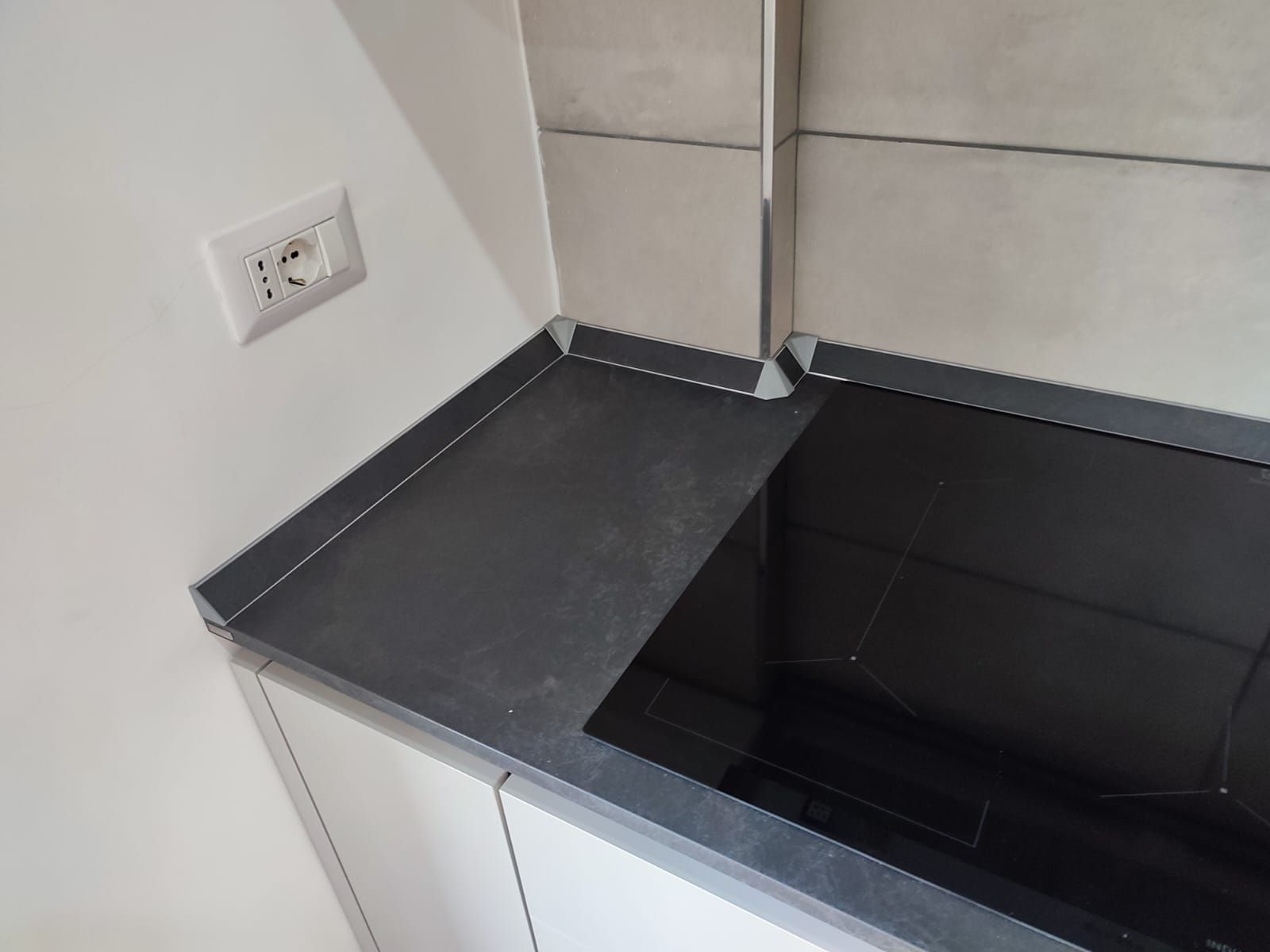 Piano di lavoro scuro accanto a un piano cottura nero, con una presa elettrica bianca sulla parete.