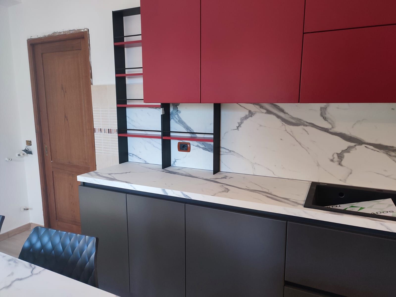 Cucina moderna con mobili rossi e grigi, ripiani in marmo e scaffali a vista.