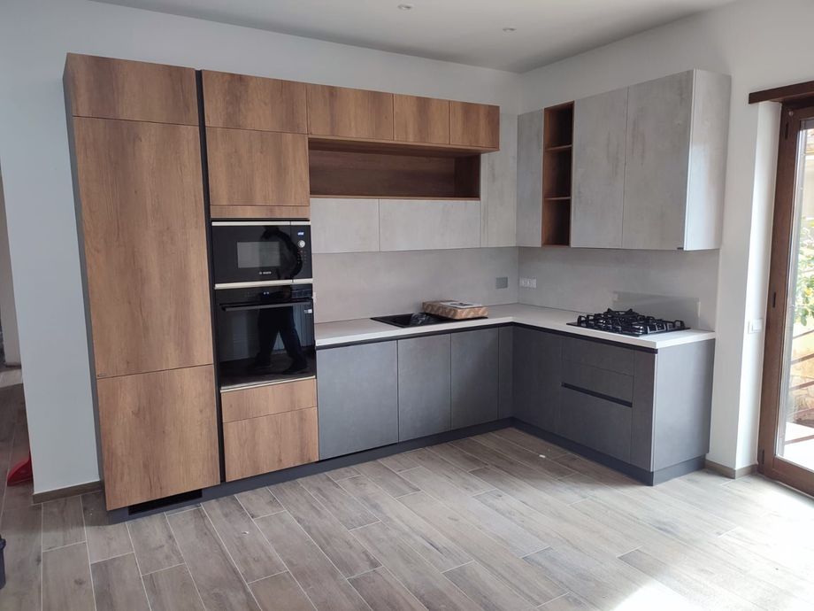 Cucina moderna con mobili in legno chiaro e grigio, forni incorporati e piano cottura.