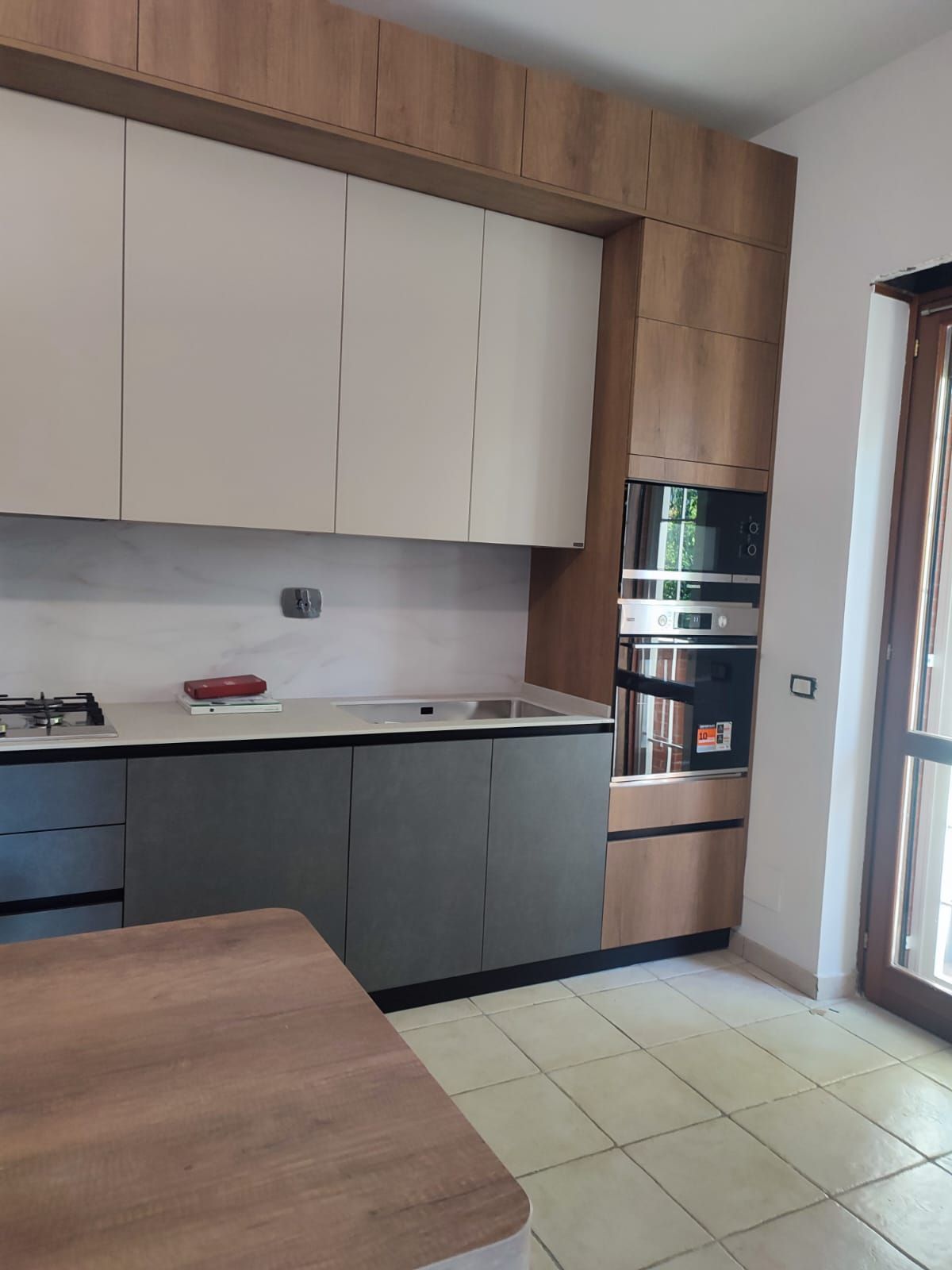 Cucina moderna con mobili grigi, bianchi e in legno, forno incorporato e tavolo in legno.