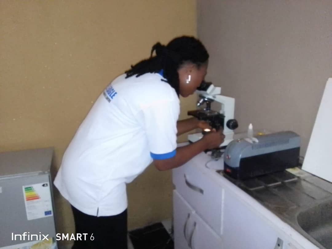 A woman looking through a microscope with a infinix smart 6 in the background