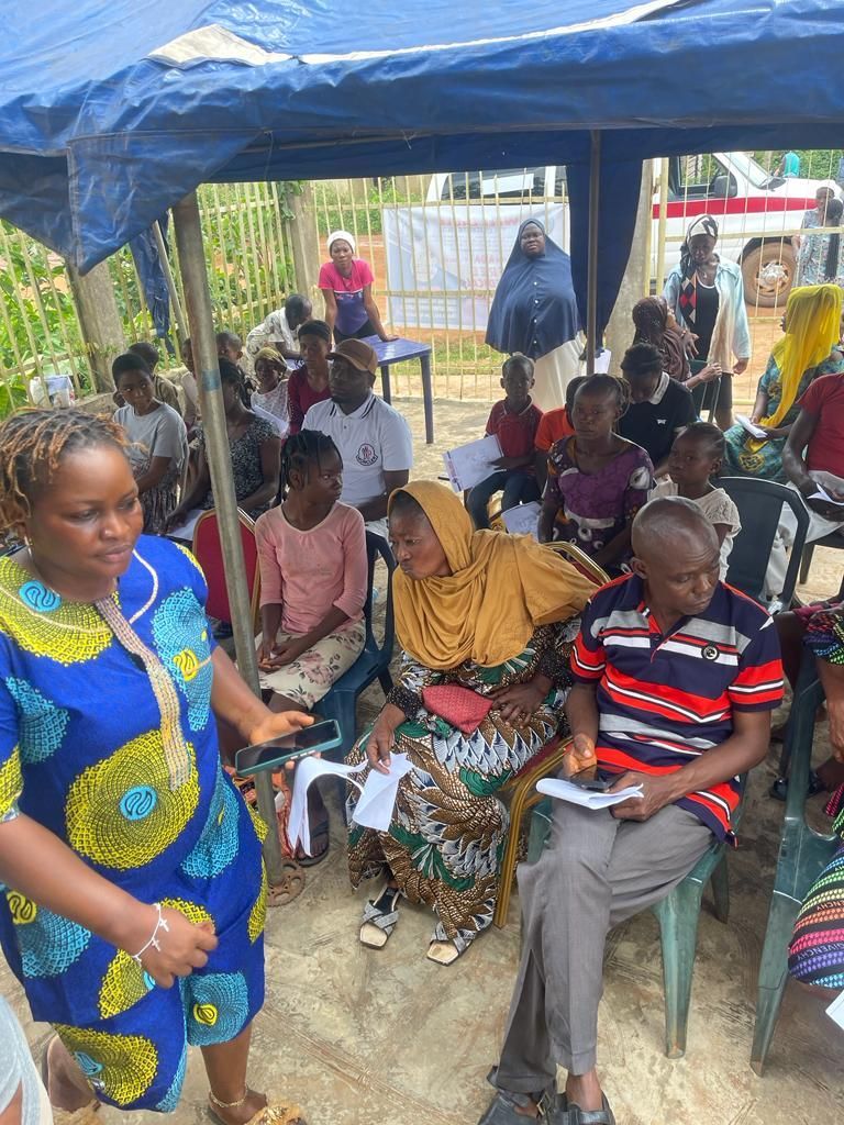 A woman is standing in front of a group of people sitting under a tent.