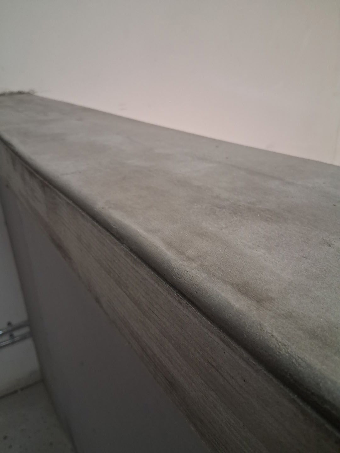 Close-up of a gray concrete surface, likely a ledge, with a rounded edge, against a white wall.