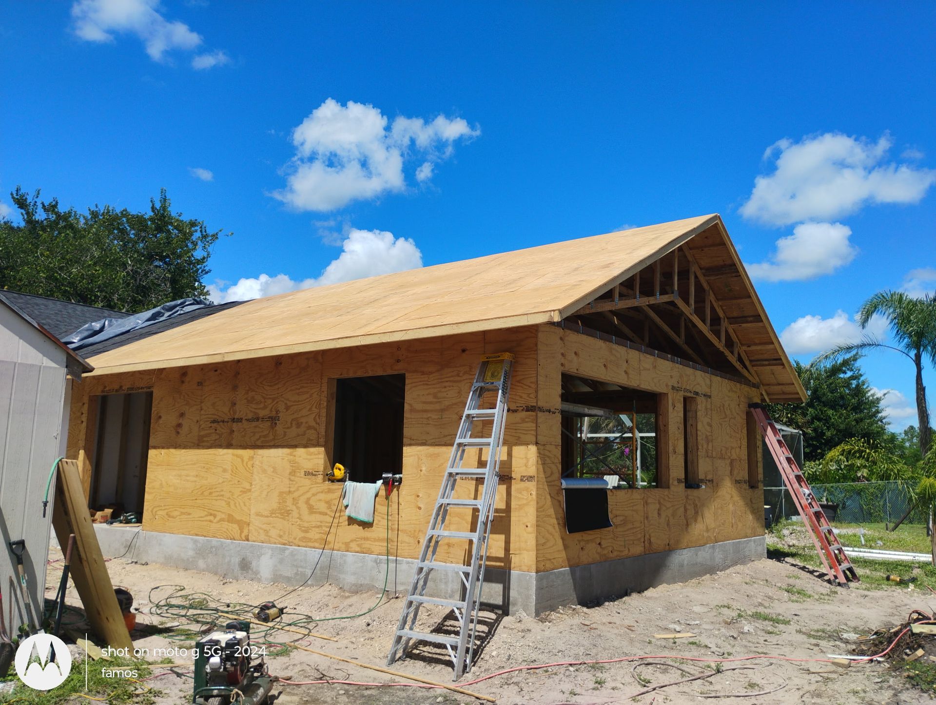 Construction of a small building, with plywood walls and roof frame, set against a bright blue sky.