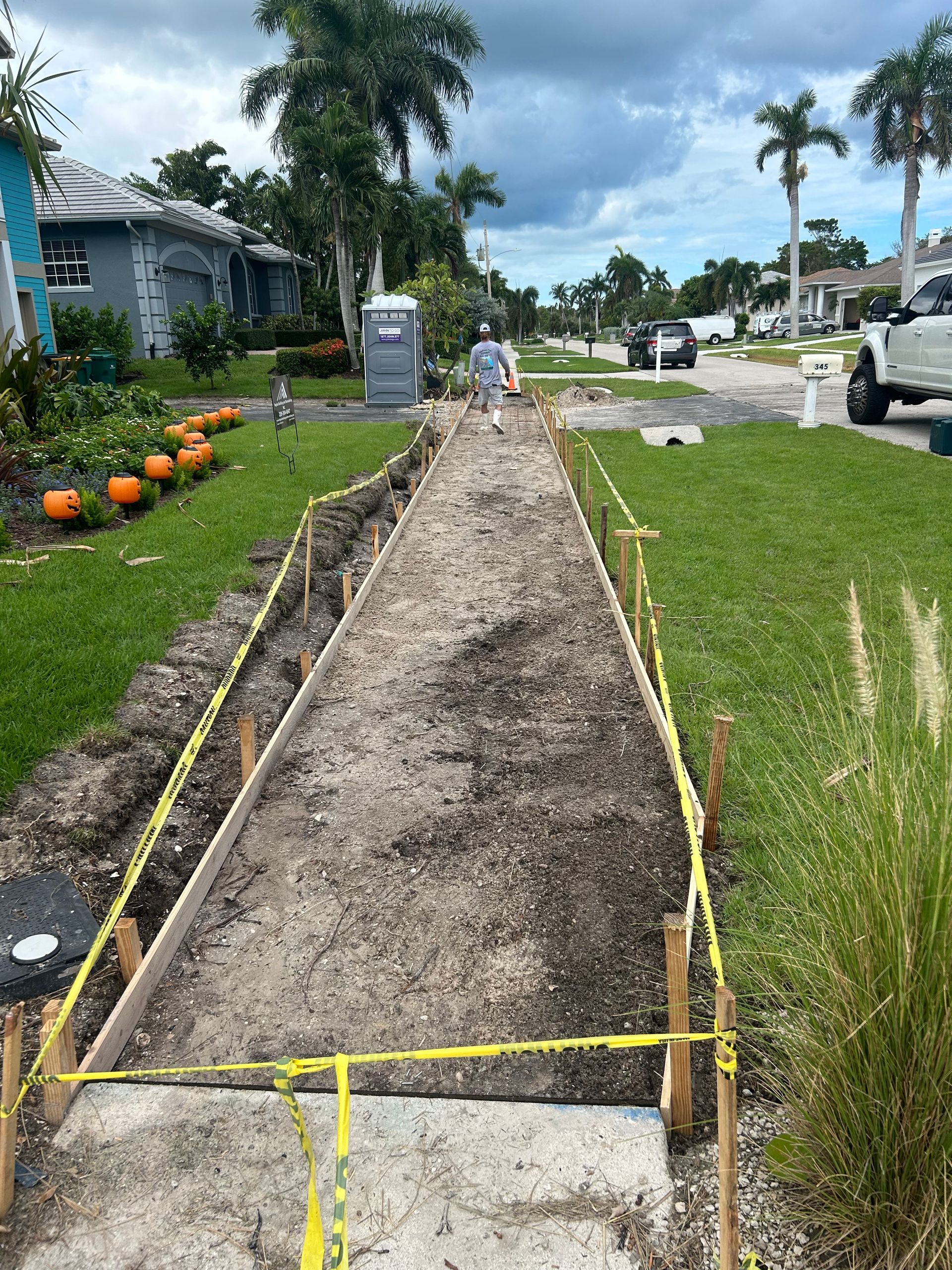 Sidewalk construction: dirt path between lawn & street, yellow caution tape, worker, portable toilet.