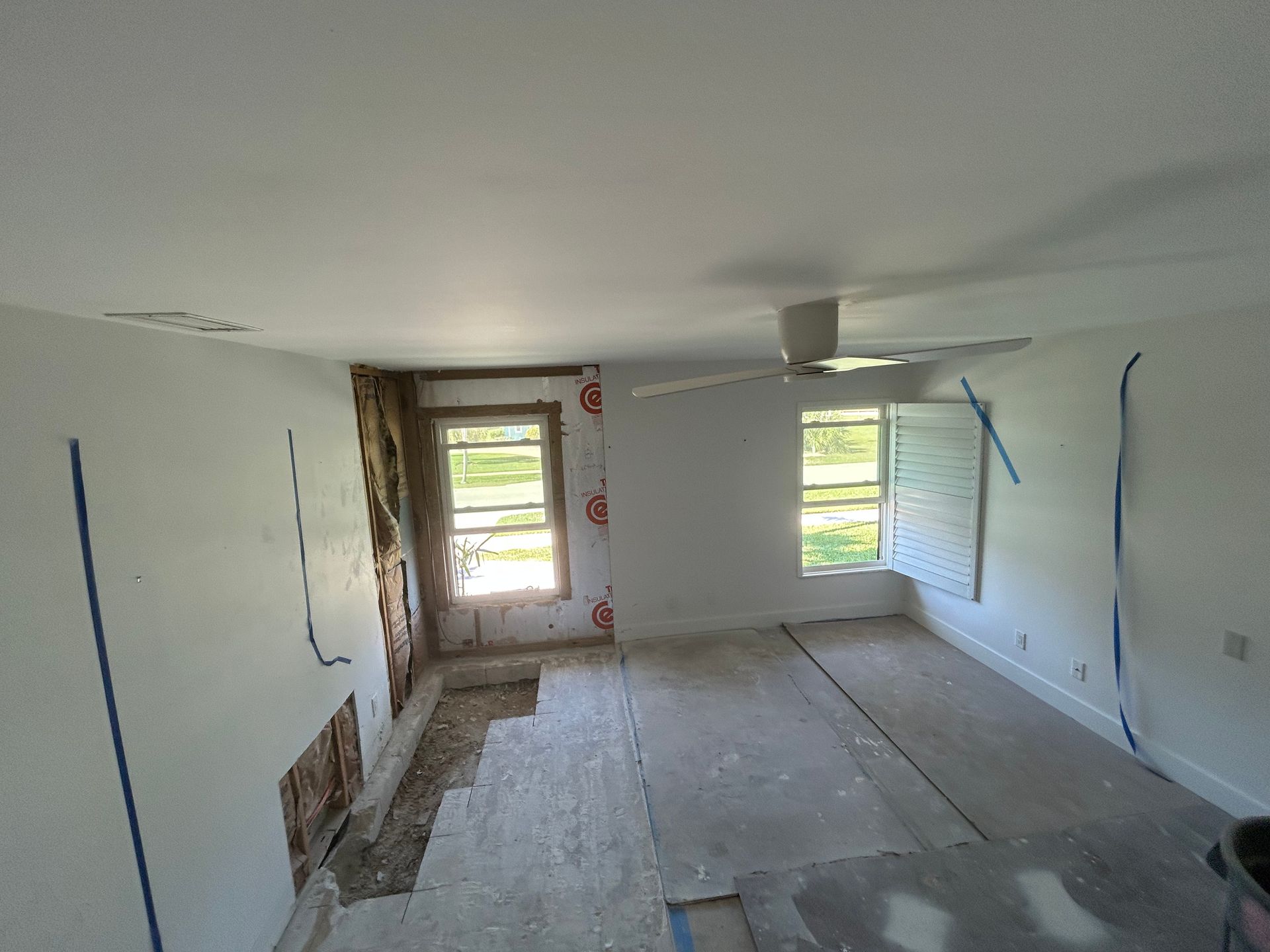 Room under renovation with exposed wall, window, and ceiling fan. White walls, blue tape, unfinished floor.