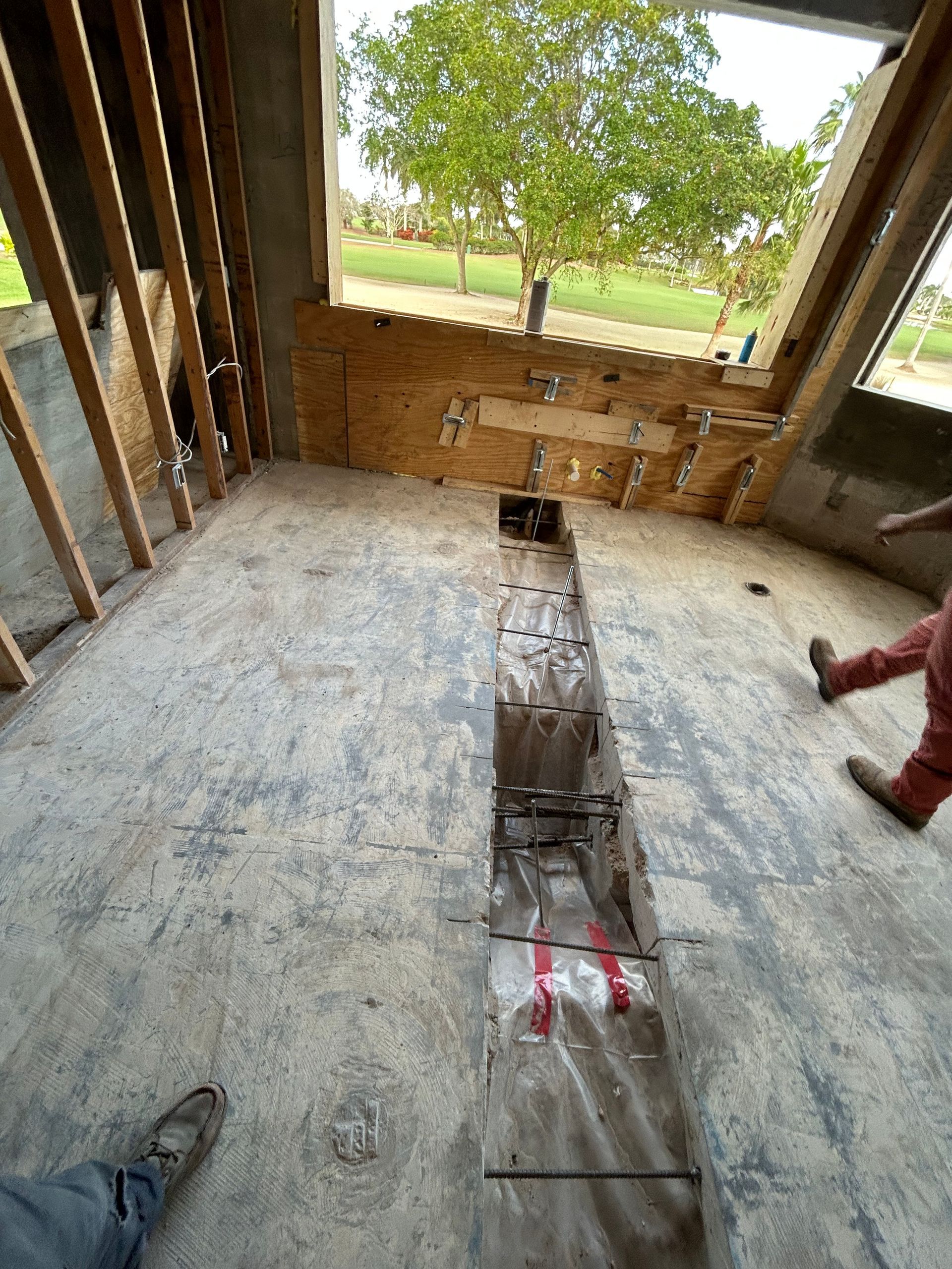 Construction site interior with exposed wooden studs, concrete floor, and window. Long trench in floor.