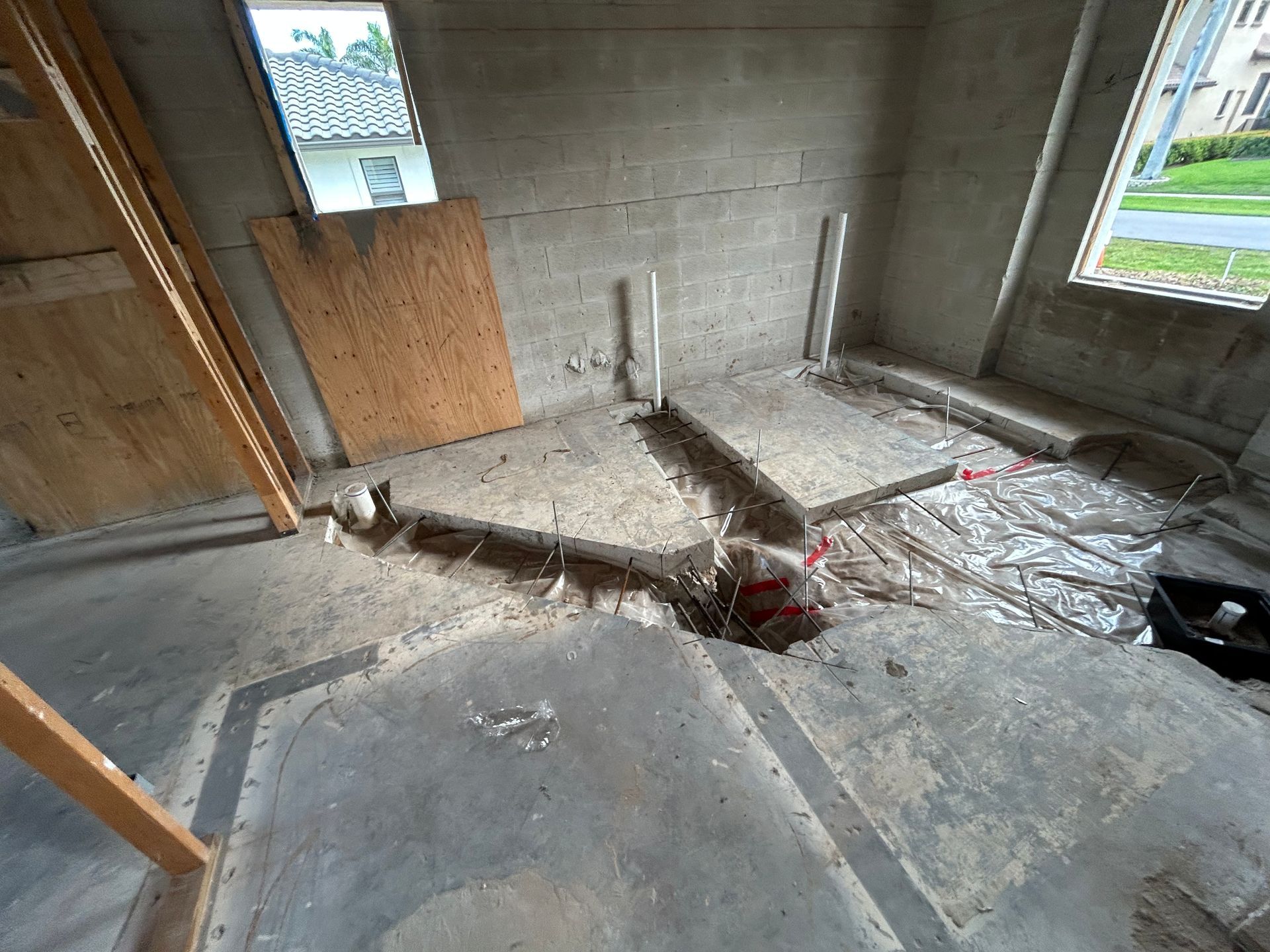 Interior room under construction with exposed pipes and wiring in concrete floor, two windows.