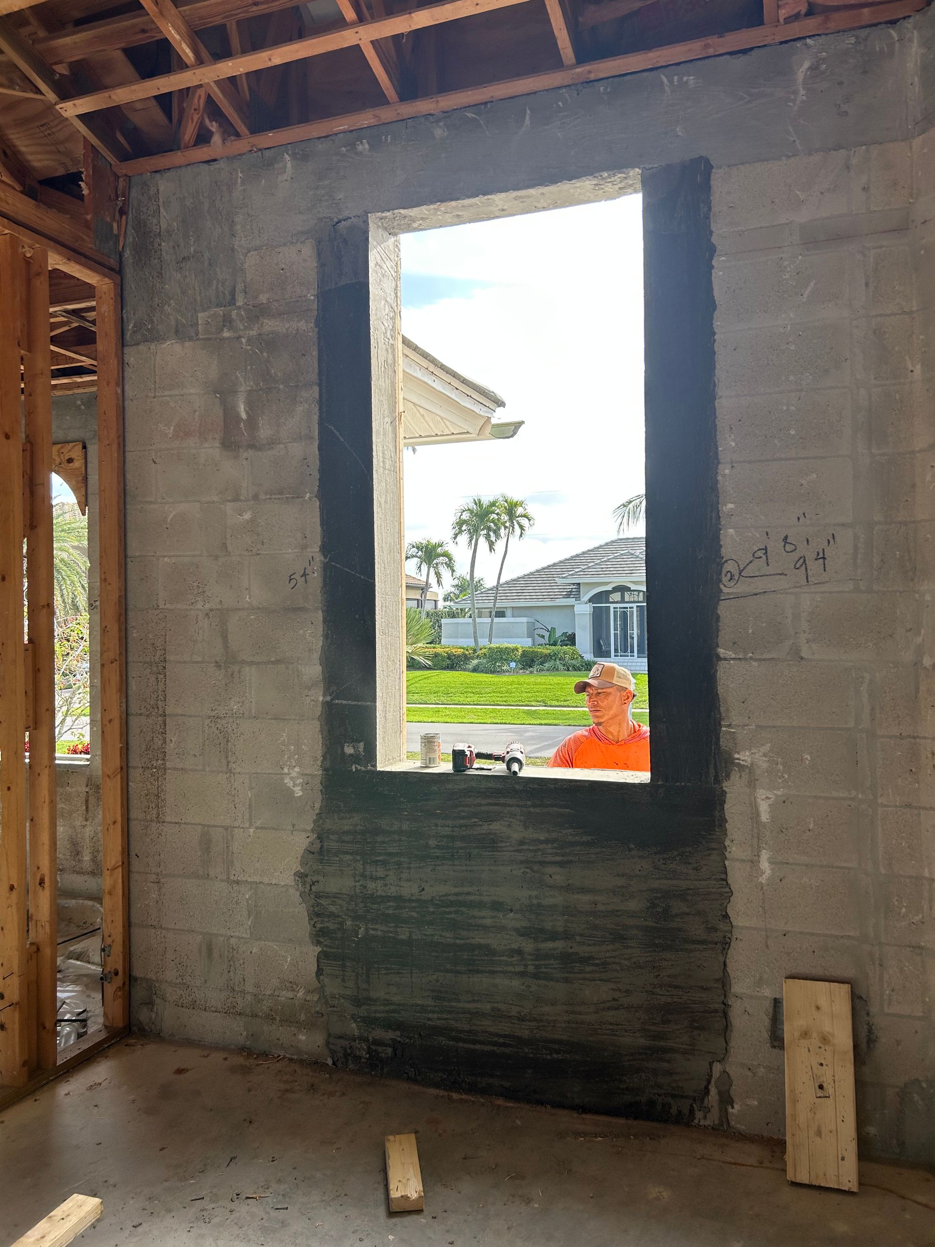 Interior view of a concrete block wall with a window opening. A person in orange vest is visible outside the window.