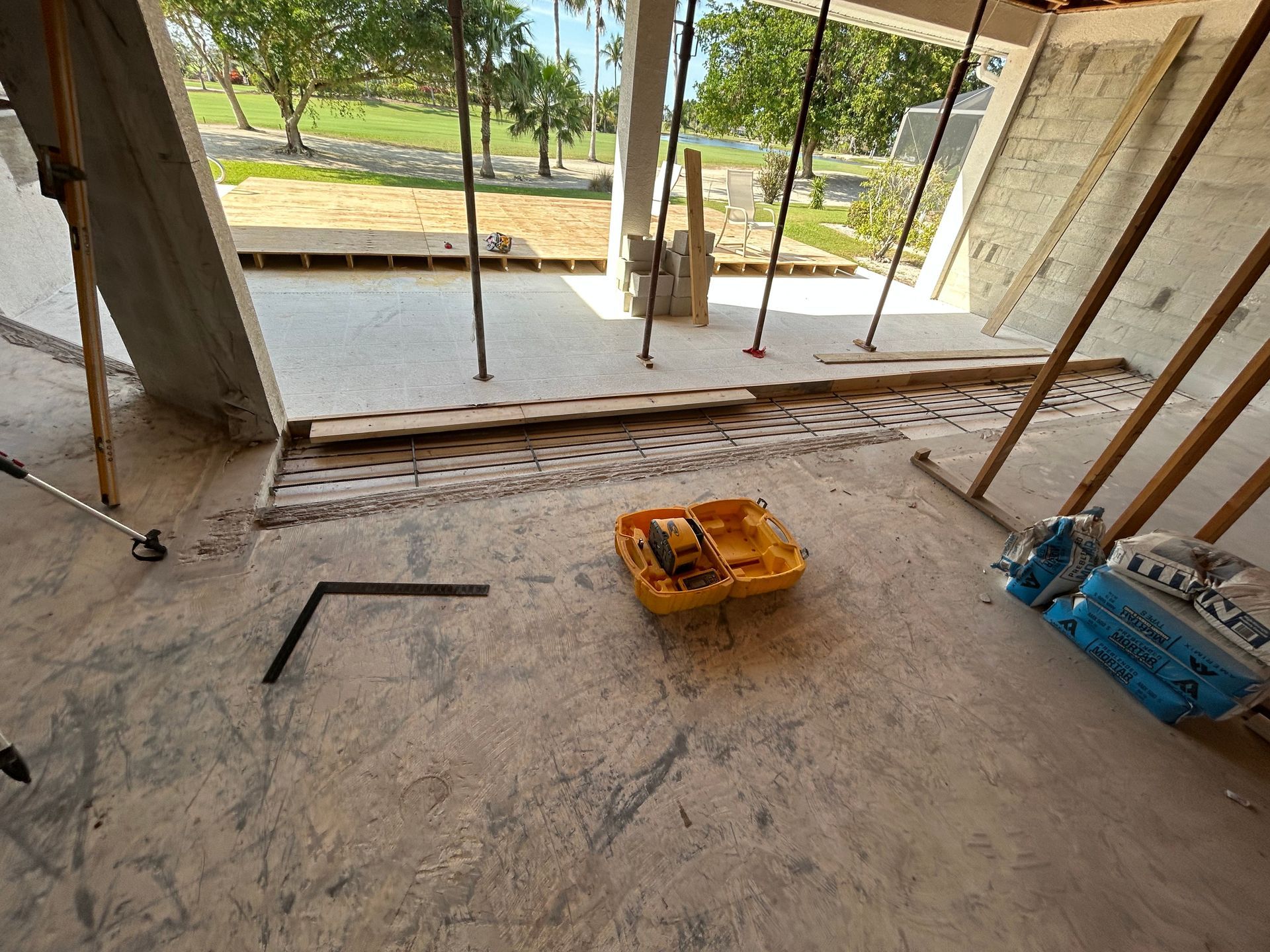 Construction site interior with concrete floor, tools, and supporting beams; view to outdoor greenery.
