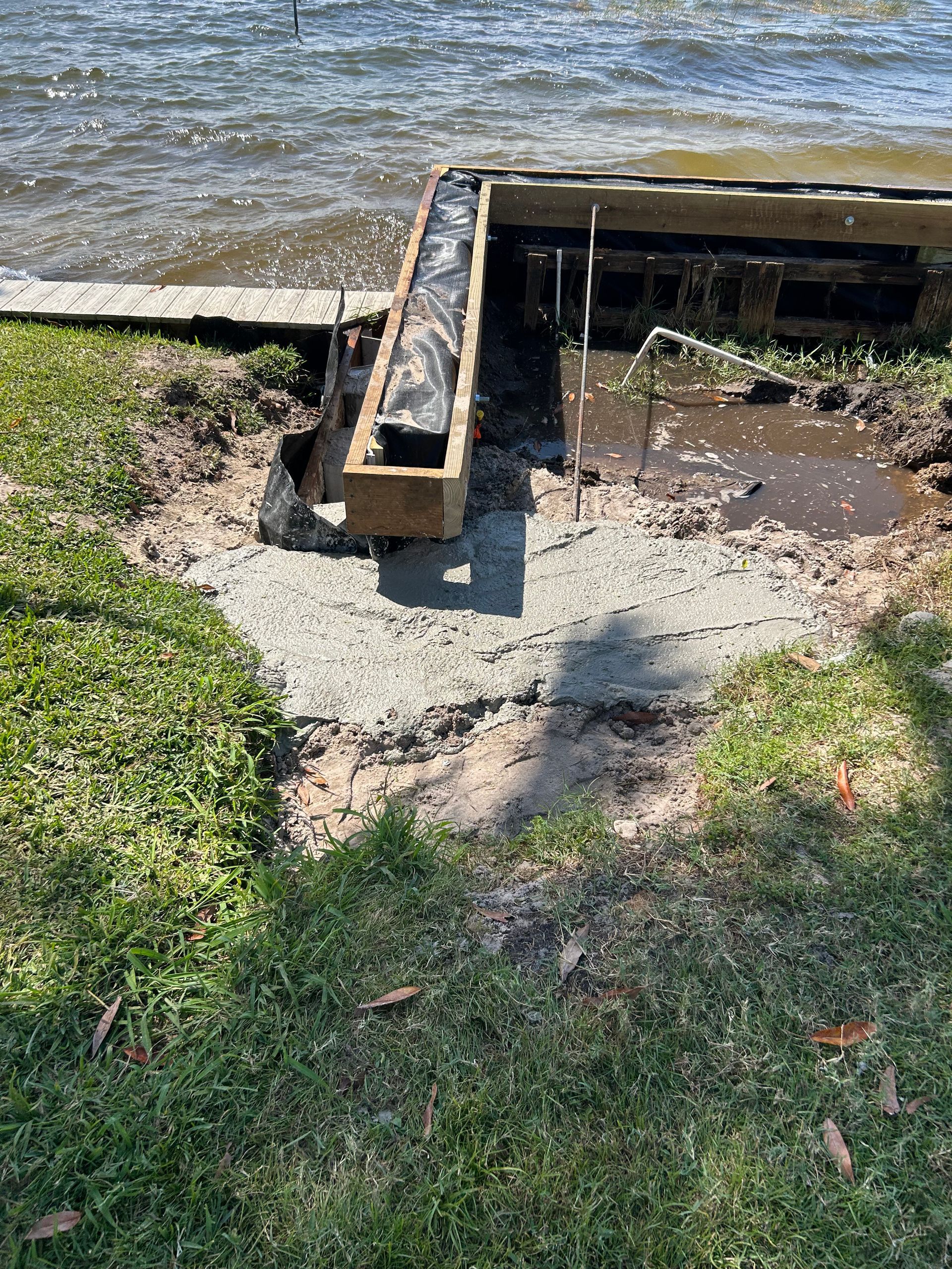 Concrete pathway and wooden structure at water's edge. Grass, water, and erosion are visible.