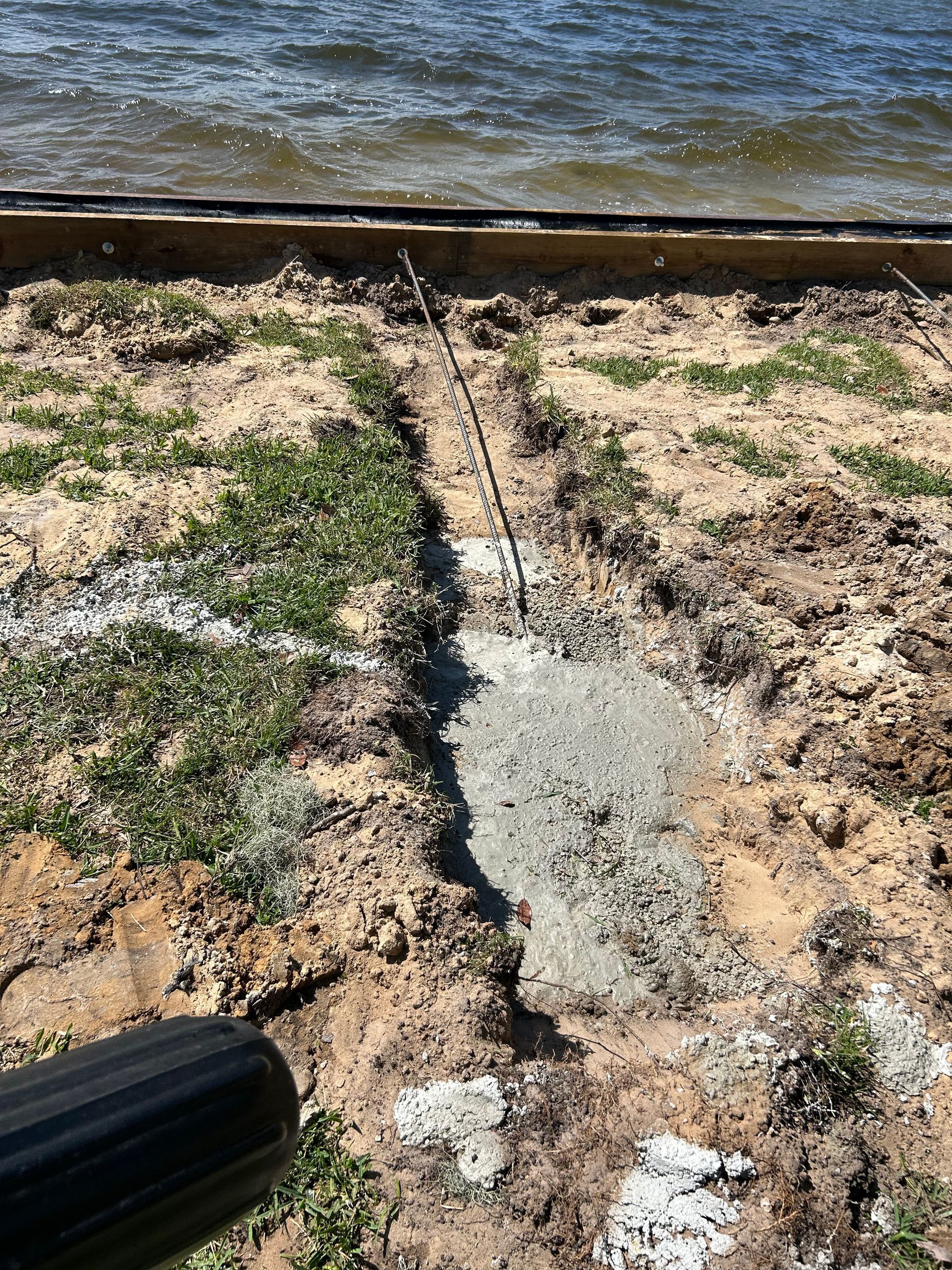 Construction site: concrete patch on sandy ground near water. A pipe and vegetation are visible.