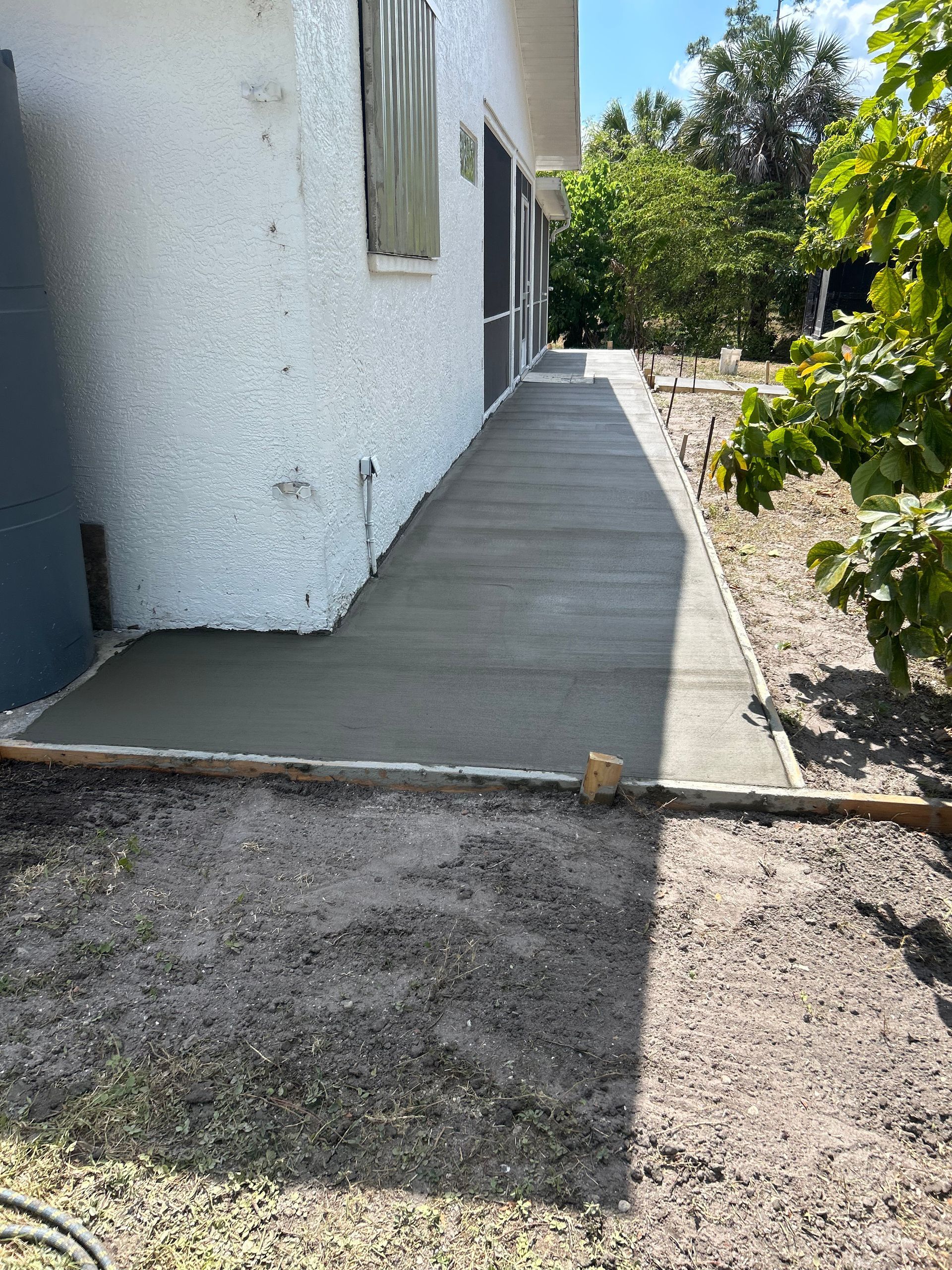Freshly poured concrete walkway alongside a white stucco building with a green landscape.