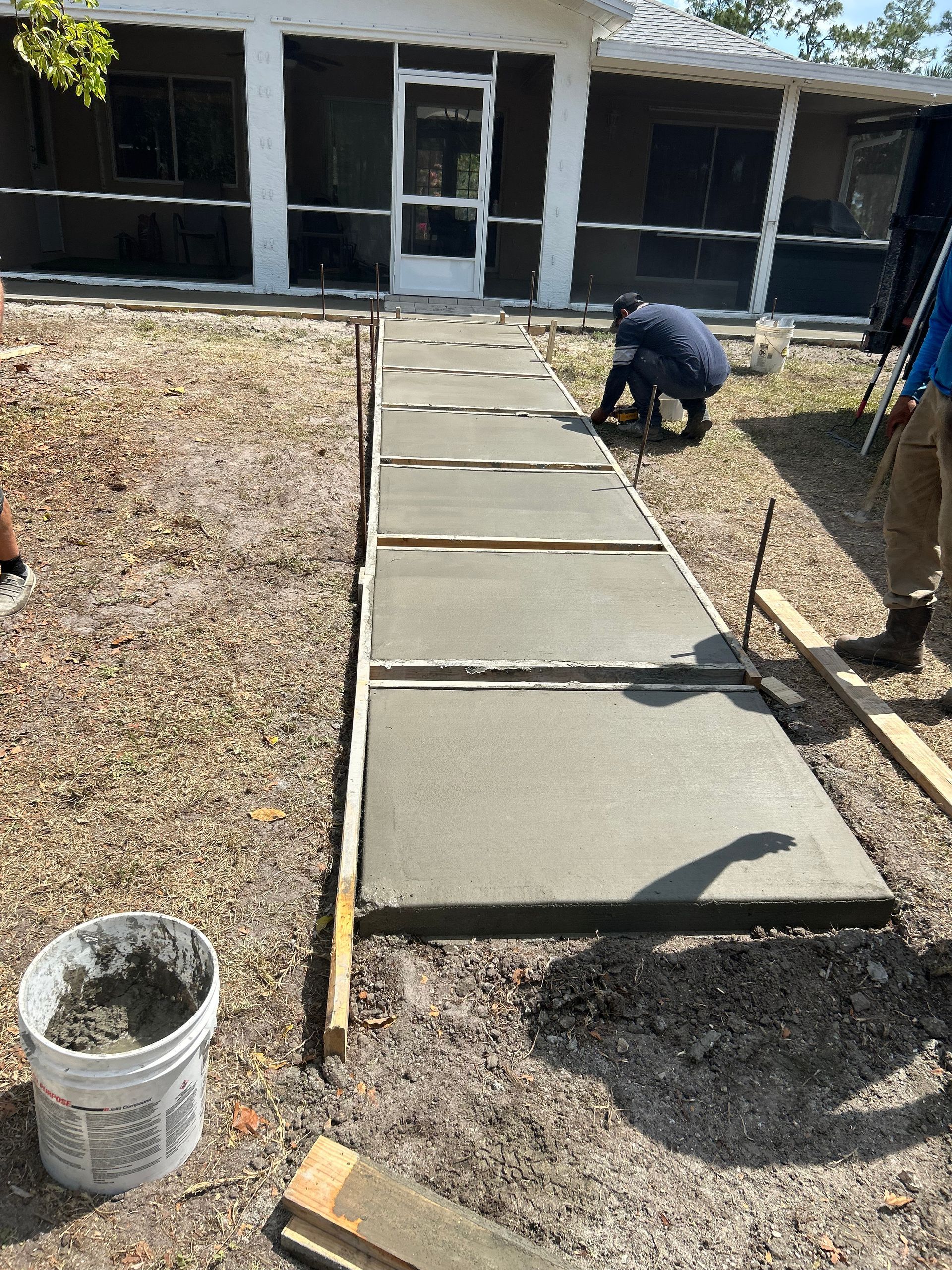 Workers pouring and smoothing concrete sidewalk, next to a building with a screen door.