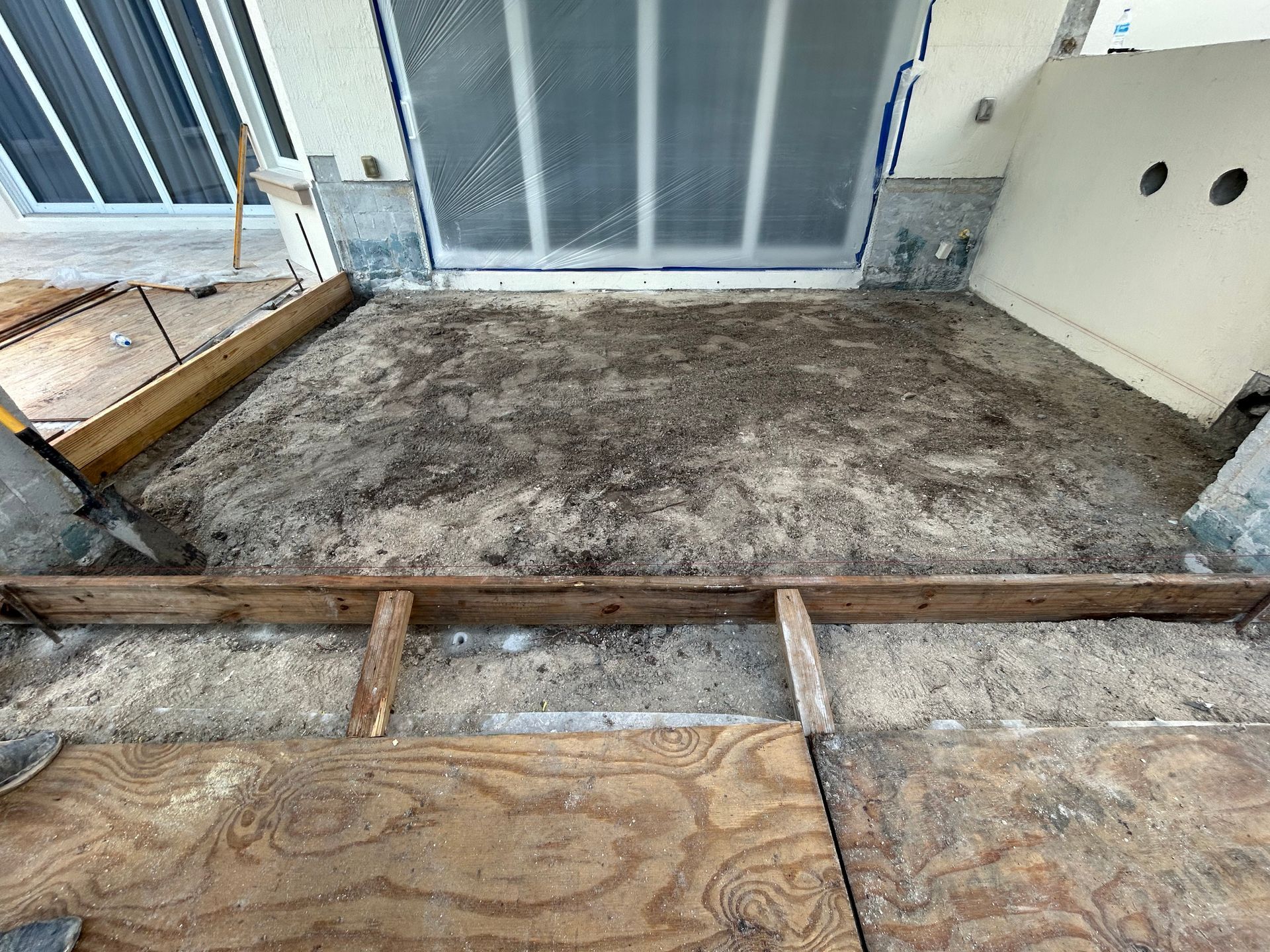 Exterior space under construction, featuring exposed subfloor, sand, wooden framing, and a sliding door.