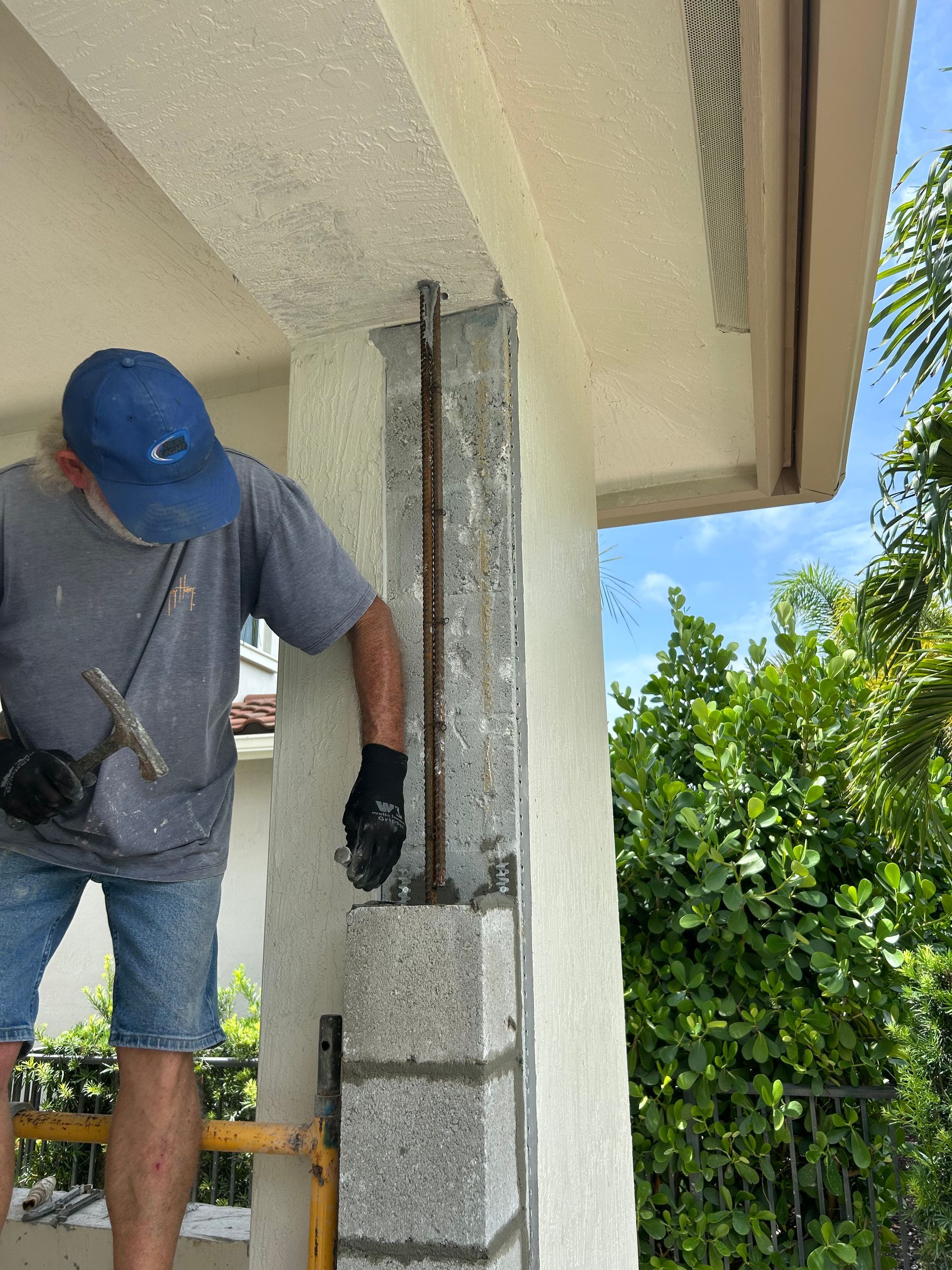 Man works on concrete block column, holding a tool. Rebar is visible. Exterior setting, daytime.