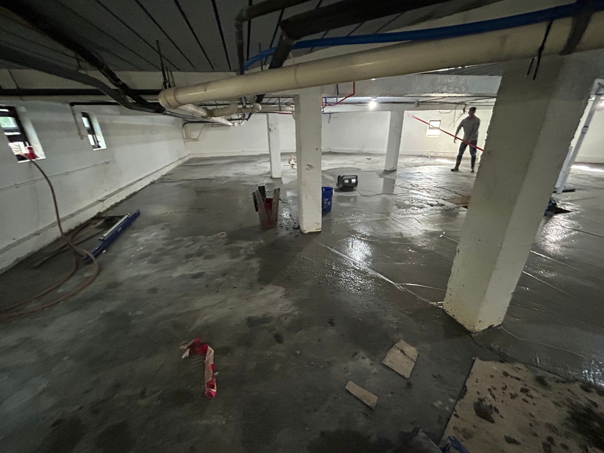 Man washing concrete floor in a basement with support pillars and pipes overhead.