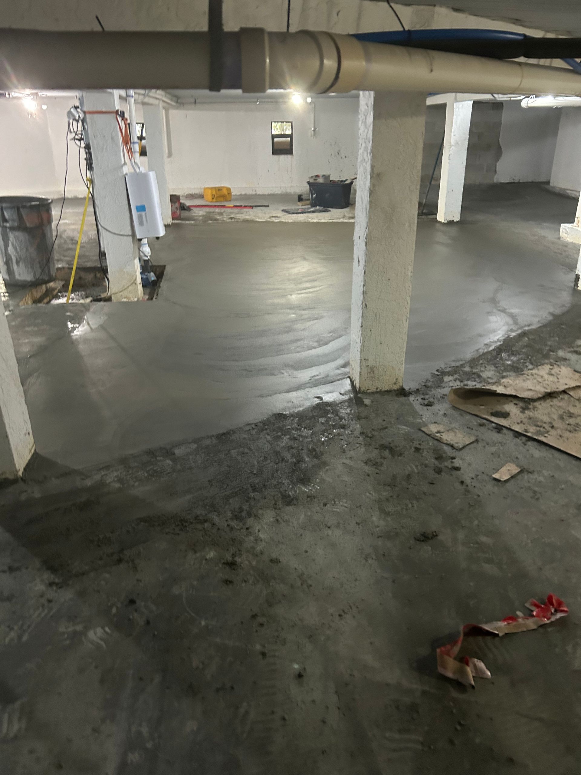 Concrete floor freshly poured in a basement, with support columns and pipes overhead.