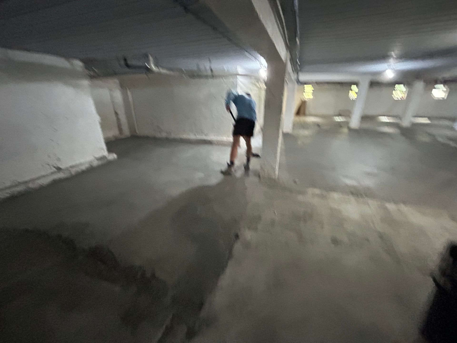 Person sweeping a concrete floor in a large, dimly lit, mostly empty room.