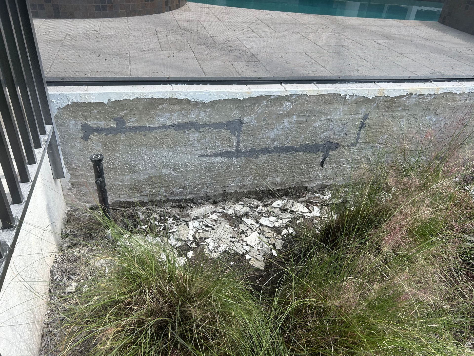 Concrete wall section next to a pool with exposed foundation, grass, and a black metal pole.