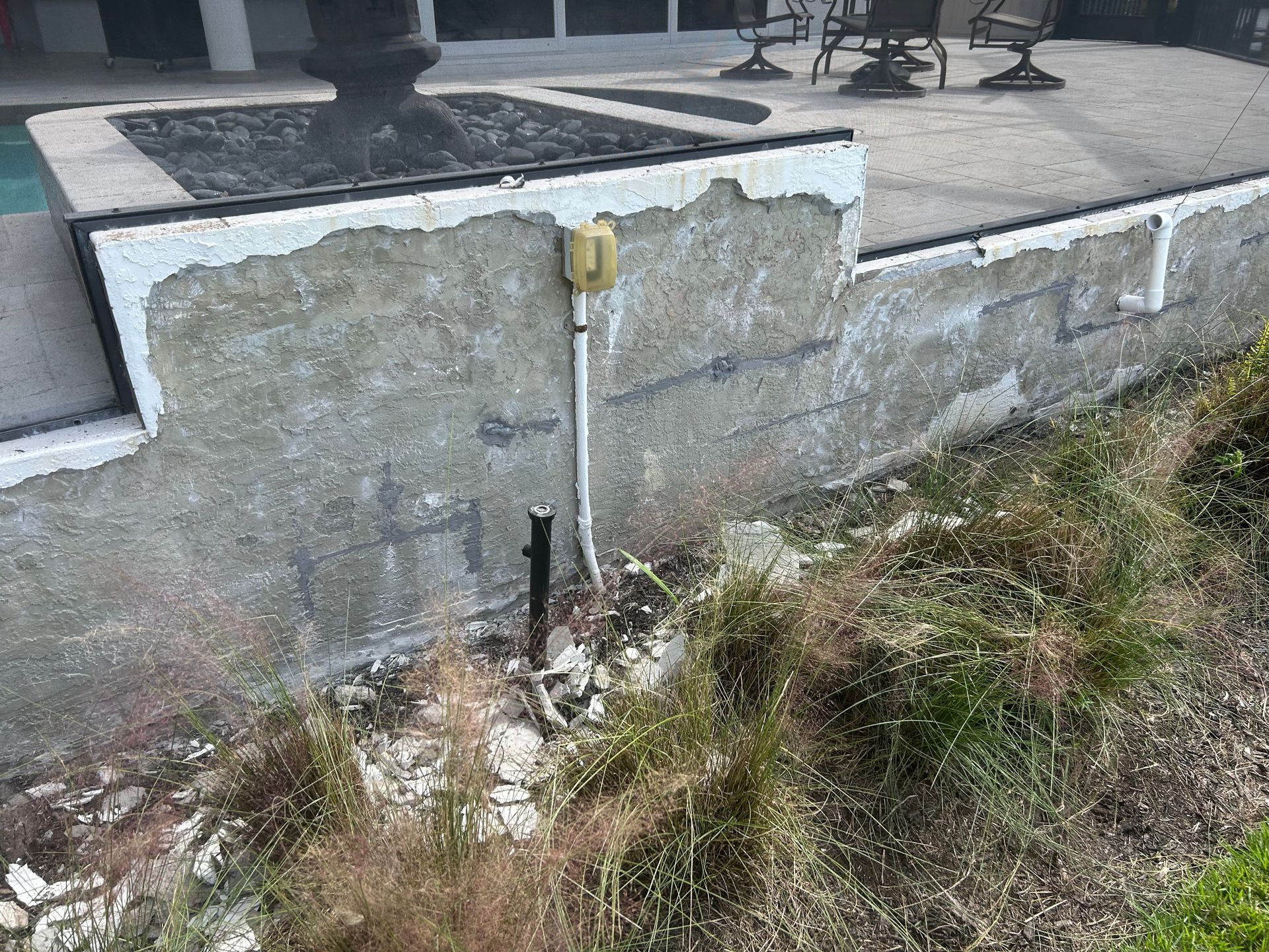 Damaged concrete wall with exposed wiring and overgrown vegetation.
