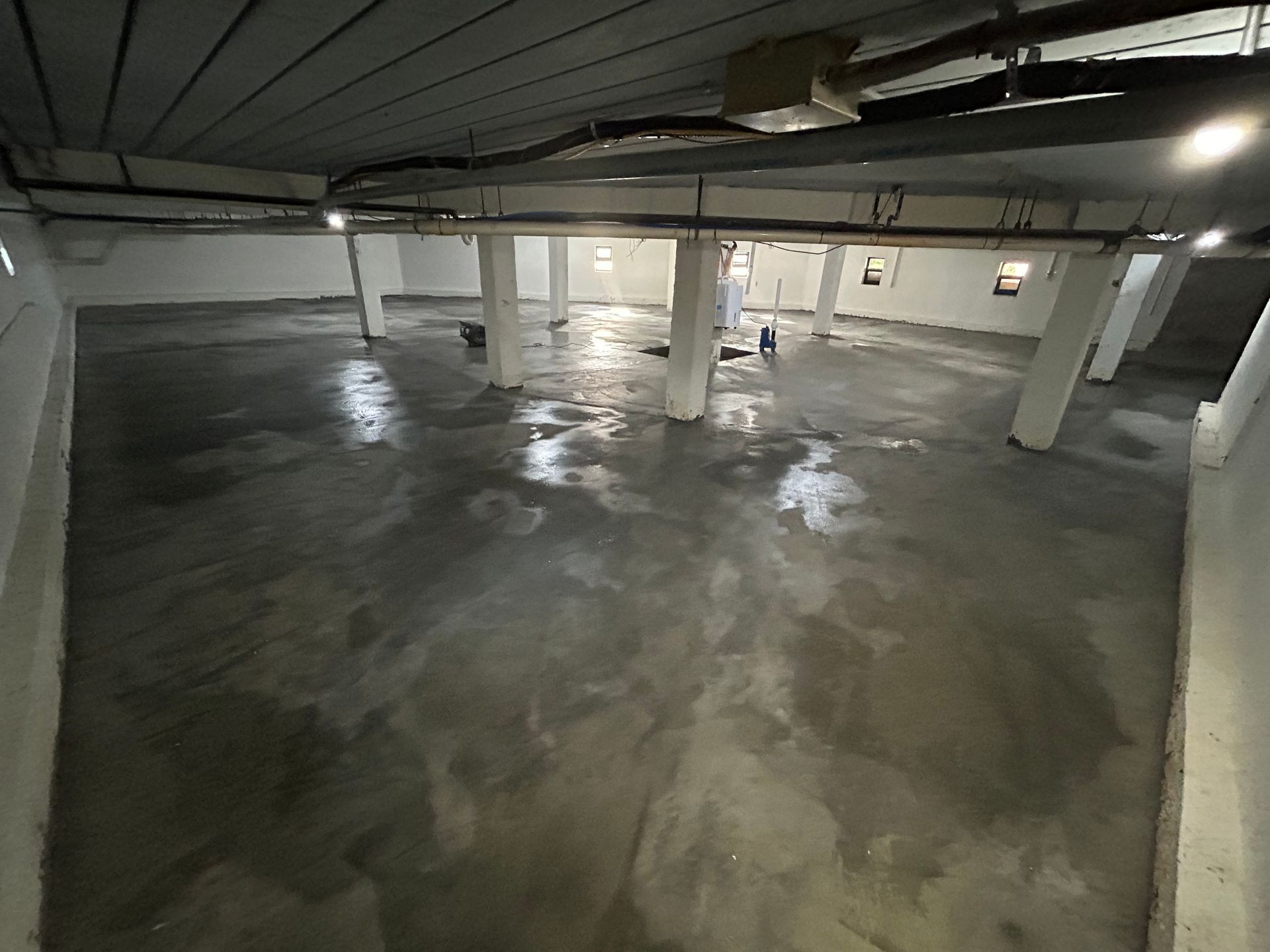 Wet, gray concrete floor in a large, empty basement with support columns and overhead pipes.