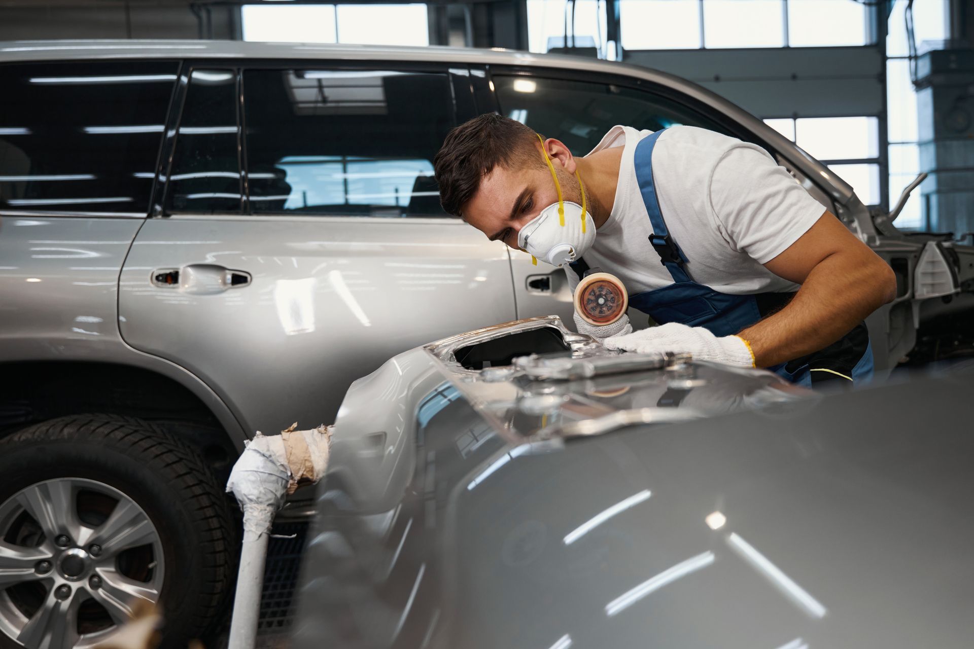 A man wearing a mask is working on a car in a garage. A man wearing a mask is working on a car in a garage.