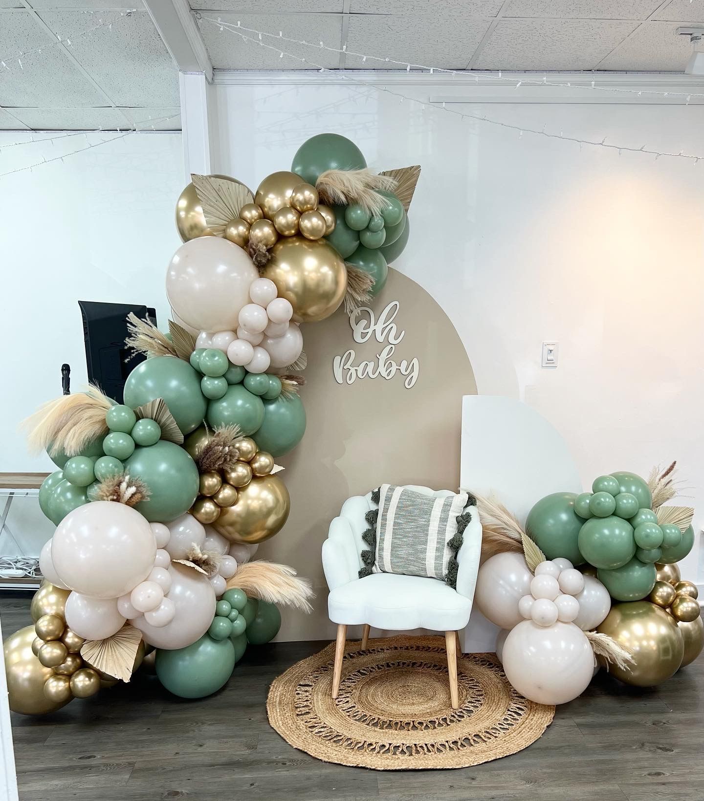 Balloon arch in sage green, gold, and cream colors with a chair and sign, for a baby shower.