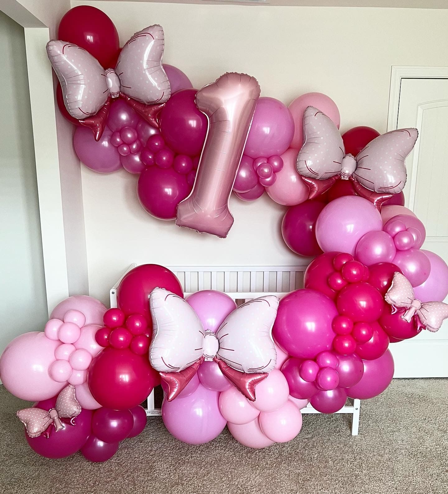 Pink and magenta balloon arch with Minnie Mouse bows and a number one, decorating a white crib.