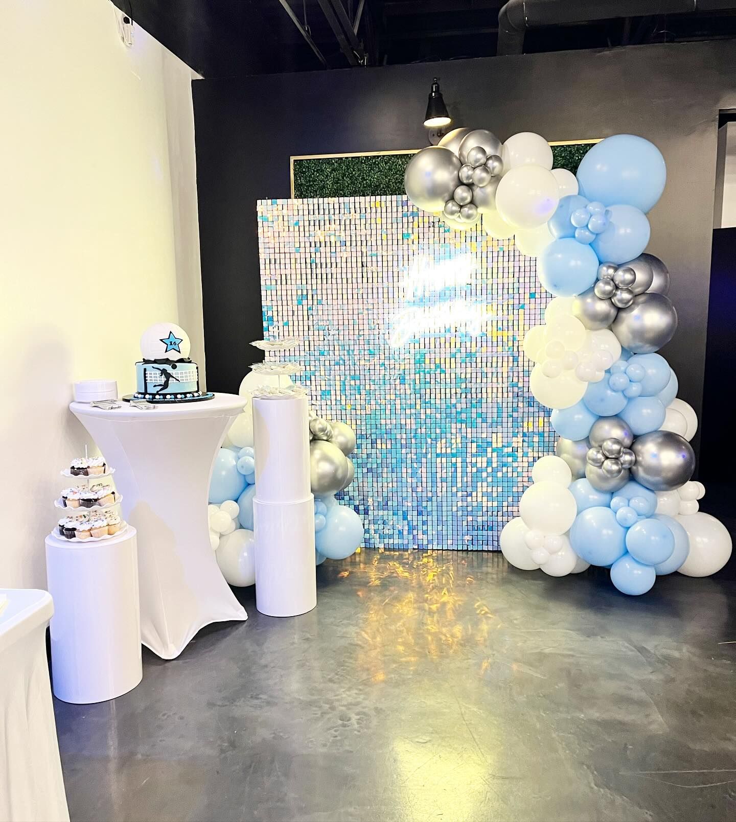 Party decorations: blue, silver, white balloon arch, cake on a table, and shimmering backdrop.