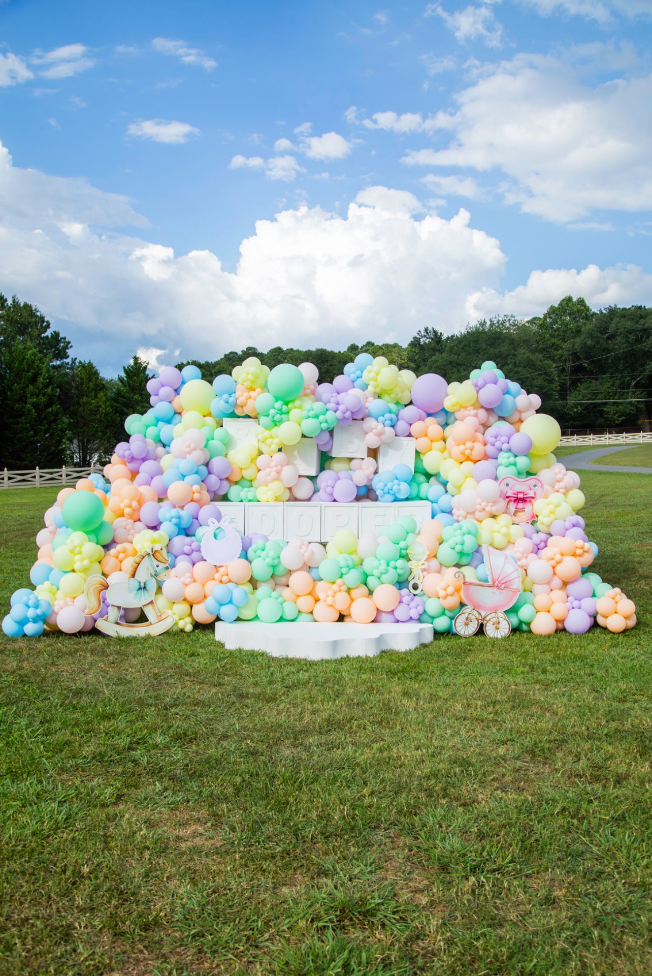 Colorful balloons layered on and around a white cinder block structure, outdoors on grass. Blue sky.